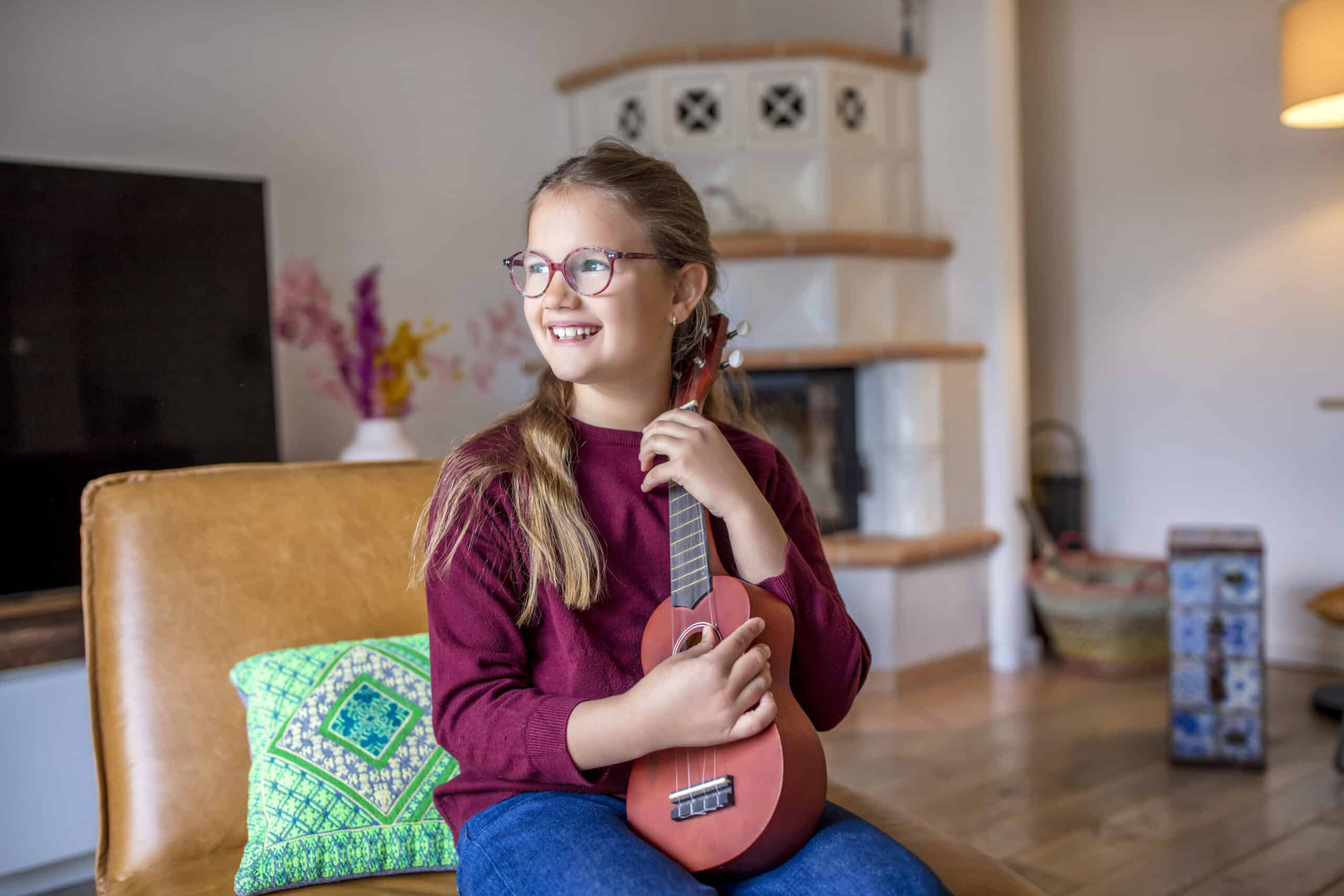 Ein junges Mädchen mit Brille und langen Haaren hält eine kleine Gitarre in der Hand, während sie auf einem Sofa sitzt. Sie trägt einen kastanienbraunen Pullover und blaue Jeans und blickt lächelnd zur Seite. Der Raum hat eine gemütliche Atmosphäre mit einem Fernseher, einem dekorativen Kissen und einem weißen Kachelofen im Hintergrund. © Fotografie Tomas Rodriguez