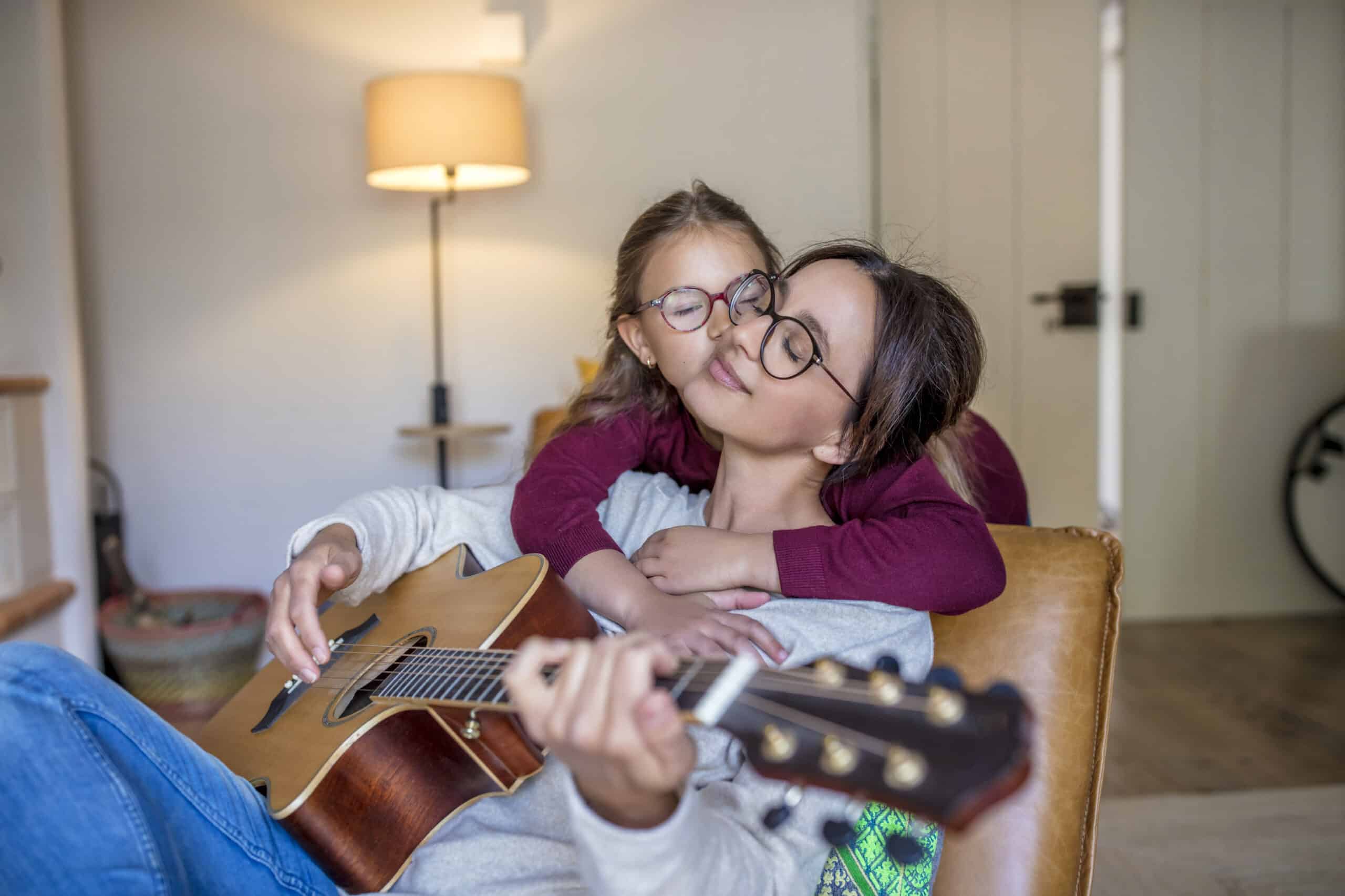 Eine Frau sitzt mit geschlossenen Augen auf einem Sofa und spielt Gitarre, während ein junges Mädchen mit Brille sie glücklich von hinten umarmt. Sie befinden sich in einem gemütlichen, gut beleuchteten Wohnzimmer mit einer Lampe und etwas Wohndekor im Hintergrund. © Fotografie Tomas Rodriguez