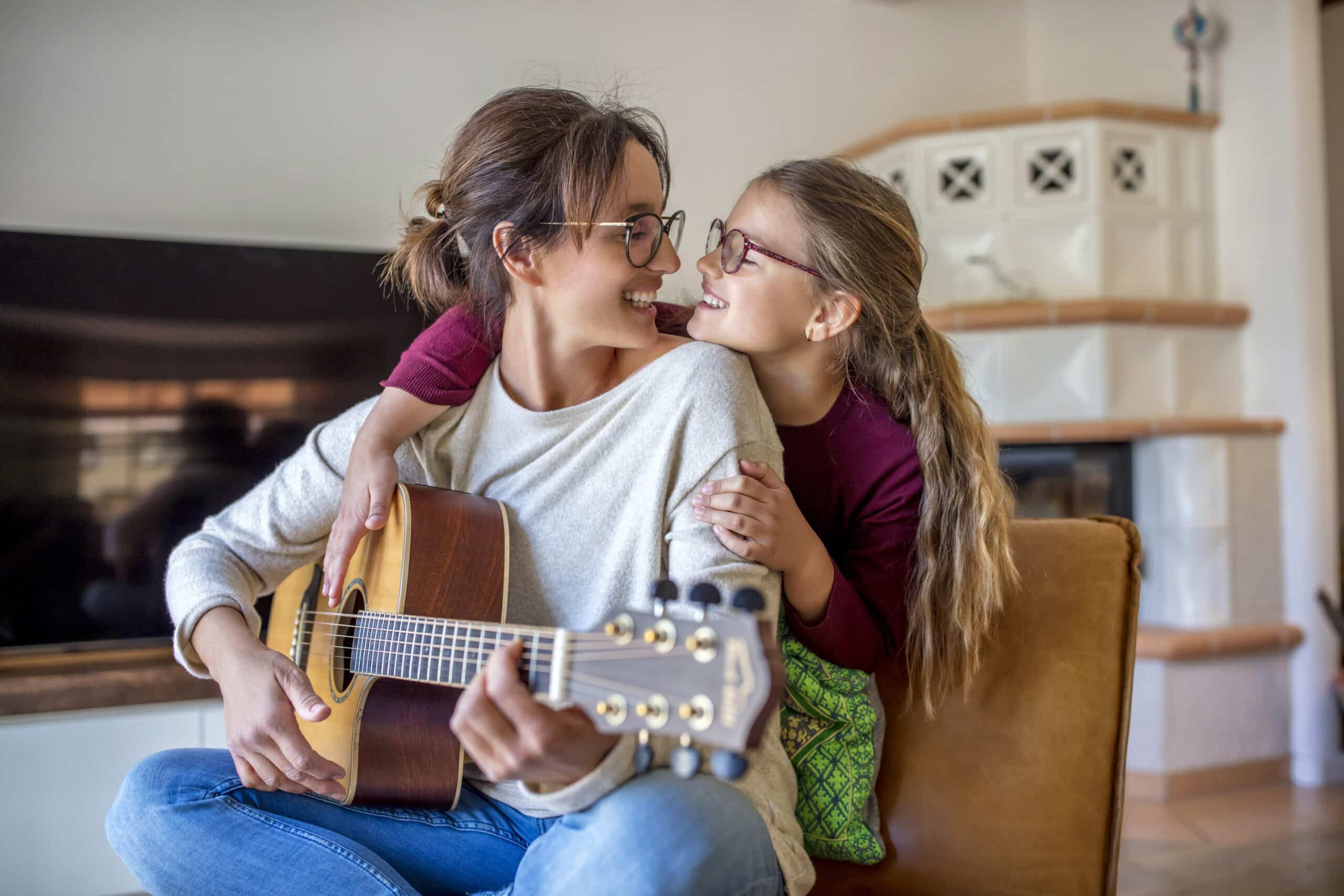 Eine Frau sitzt auf einem Stuhl und spielt Akustikgitarre, während ein junges Mädchen sie von hinten umarmt. Beide lächeln und tragen Brillen. Sie befinden sich in einem gemütlichen Wohnzimmer mit einem Kamin und einem Fernseher im Hintergrund. © Fotografie Tomas Rodriguez