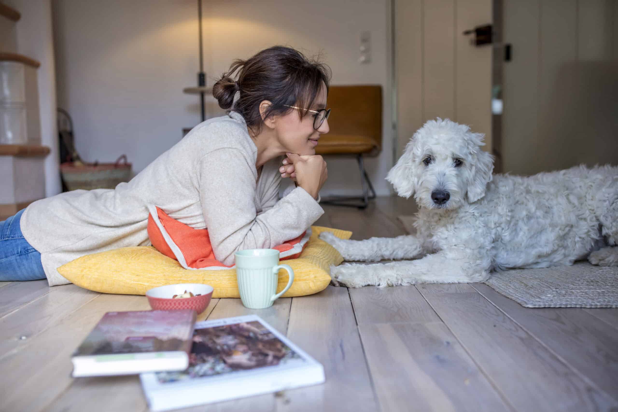 Eine Person mit Brille liegt auf dem Boden, lehnt sich auf ein orange-gelbes Kissen und blickt auf einen flauschigen weißen Hund. Eine Kaffeetasse und zwei offene Bücher liegen daneben auf dem Holzboden, im Hintergrund ist eine entspannte häusliche Umgebung zu sehen. © Fotografie Tomas Rodriguez