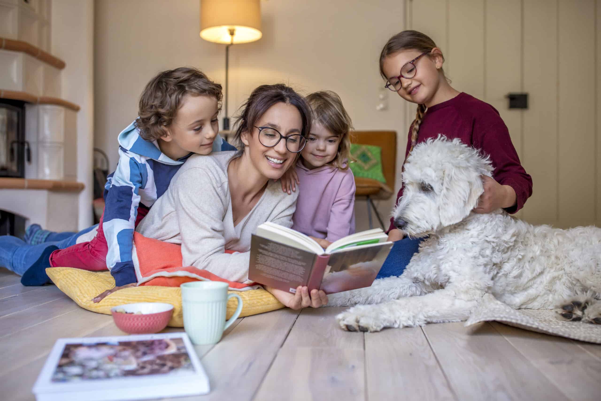 Eine Frau mit Brille sitzt auf dem Boden und liest drei Kindern, die aufmerksam um sie versammelt sind, ein Buch vor. Daneben liegt ein großer weißer Hund, den ein Kind streichelt. Im Vordergrund stehen ein Becher und eine Schüssel auf dem Boden. © Fotografie Tomas Rodriguez
