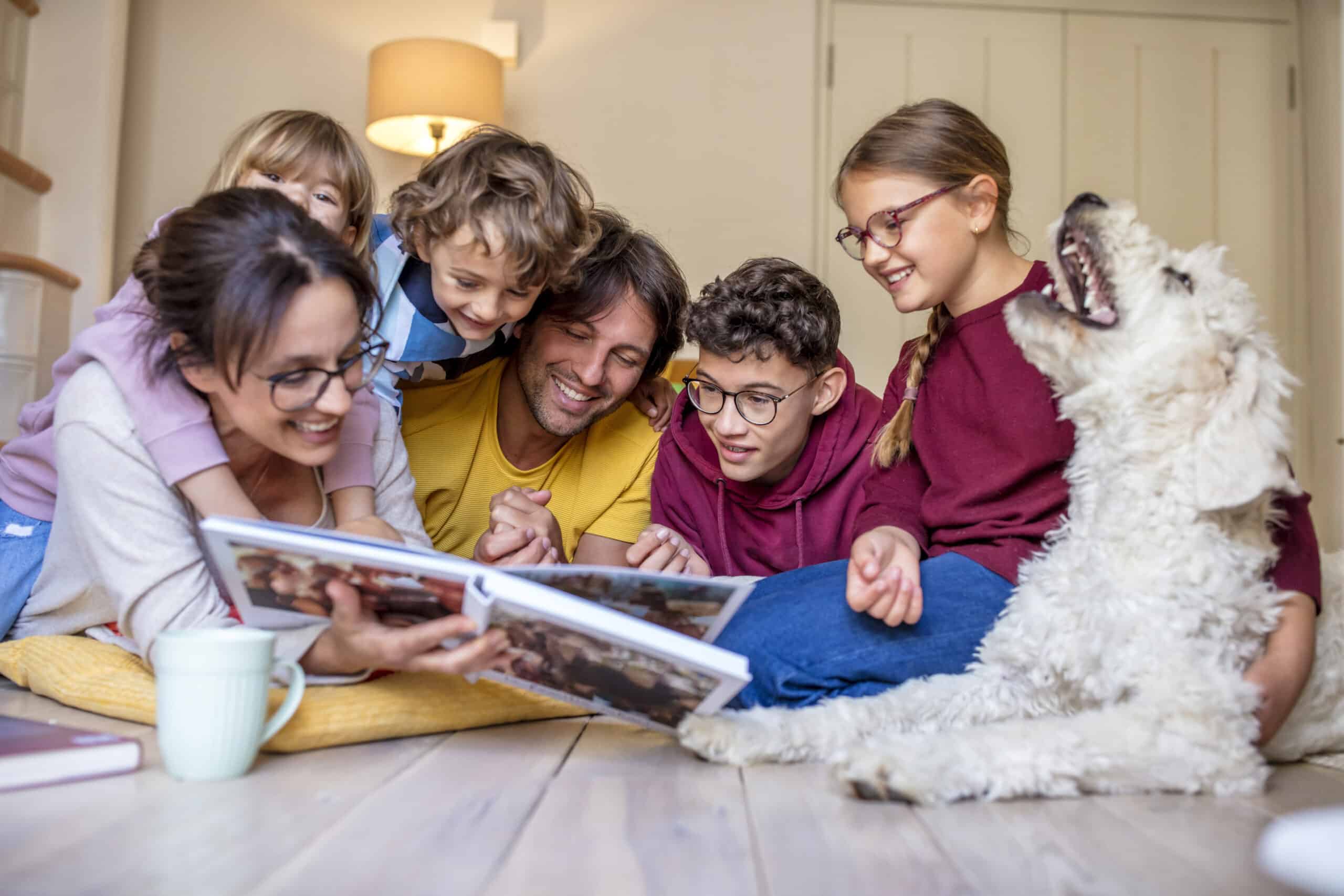 Eine sechsköpfige Familie, darunter drei Kinder, ein Teenager und zwei Erwachsene, sitzt auf dem Boden eines gemütlichen Zimmers und schaut sich glücklich ein Fotoalbum an. Ein flauschiger Hund sitzt neben ihnen und trägt zur fröhlichen Atmosphäre bei. Im Vordergrund steht eine weiße Kaffeetasse auf dem Boden. © Fotografie Tomas Rodriguez