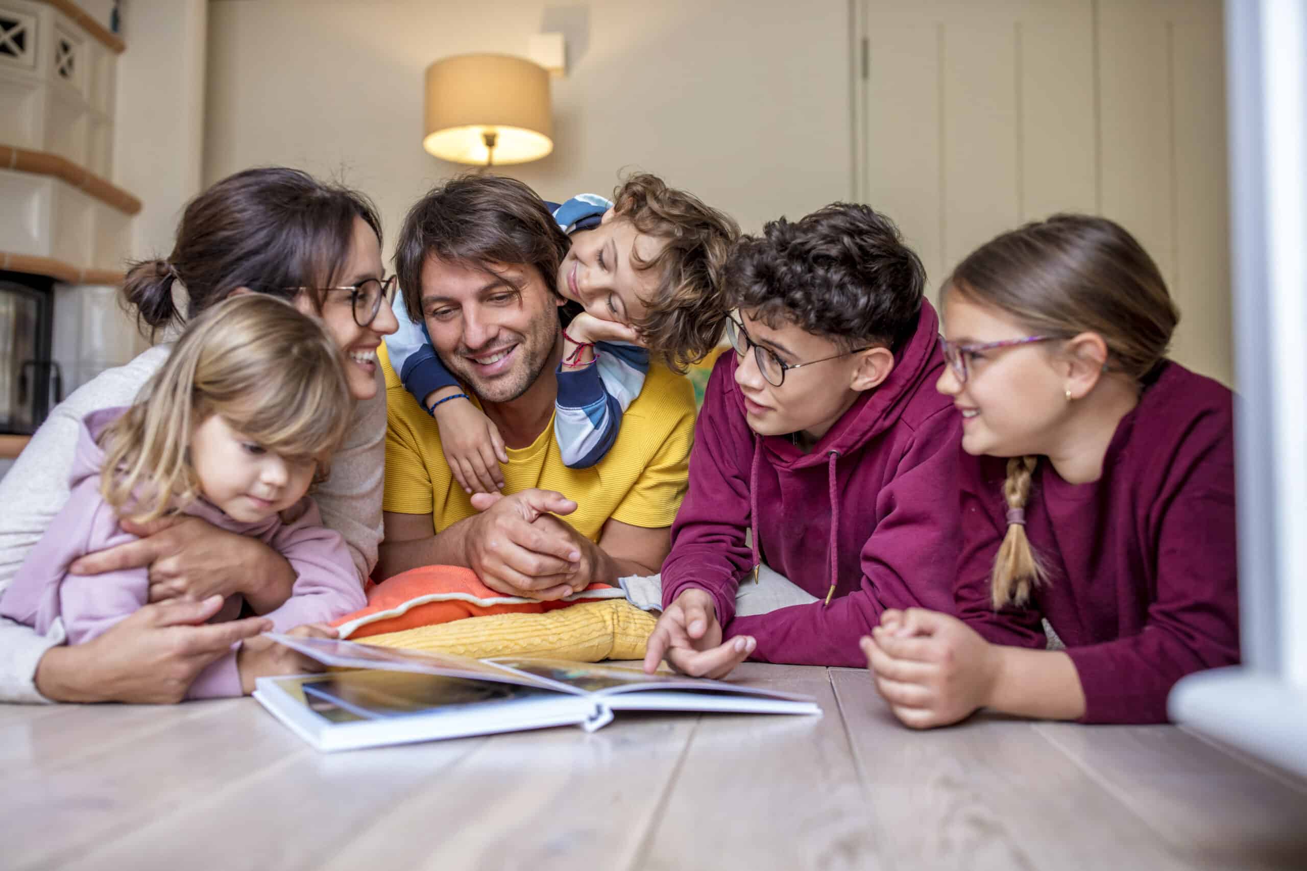 Eine Familie mit zwei Erwachsenen und vier Kindern liegt lächelnd auf dem Boden und schaut in ein offenes Buch. Die Erwachsenen und Kinder tragen Freizeitkleidung, und ein Kind ruht auf dem Rücken des Vaters. Im Hintergrund sind eine Lampe und Regale zu sehen. © Fotografie Tomas Rodriguez