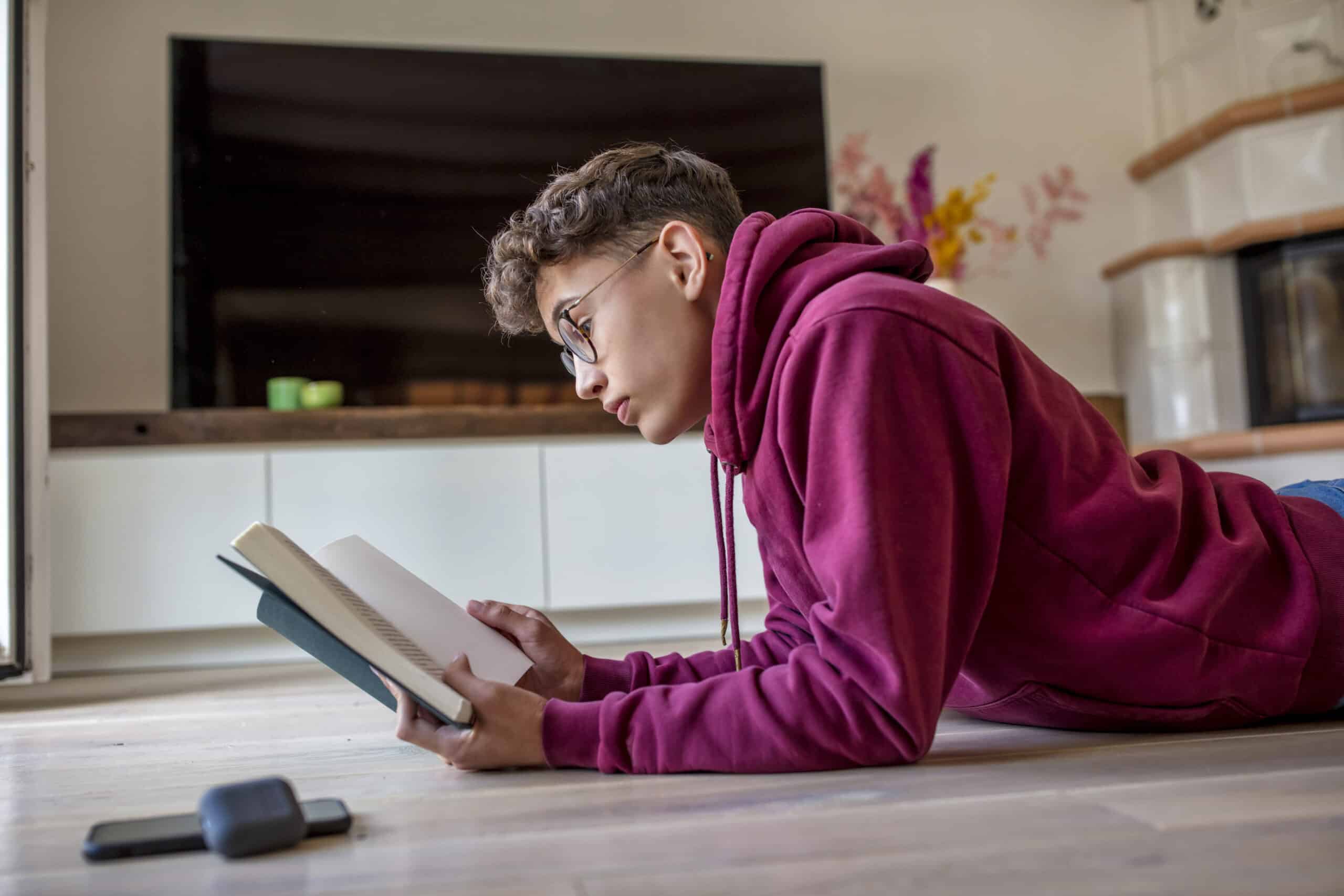 Eine Person mit Brille und kastanienbraunem Kapuzenpullover liegt auf dem Boden und liest ein Buch in einem modernen Wohnzimmer. Im Hintergrund sind ein Fernseher und eine Vase mit Blumen zu sehen, und in der Nähe steht ein Koffer auf dem Boden. © Fotografie Tomas Rodriguez