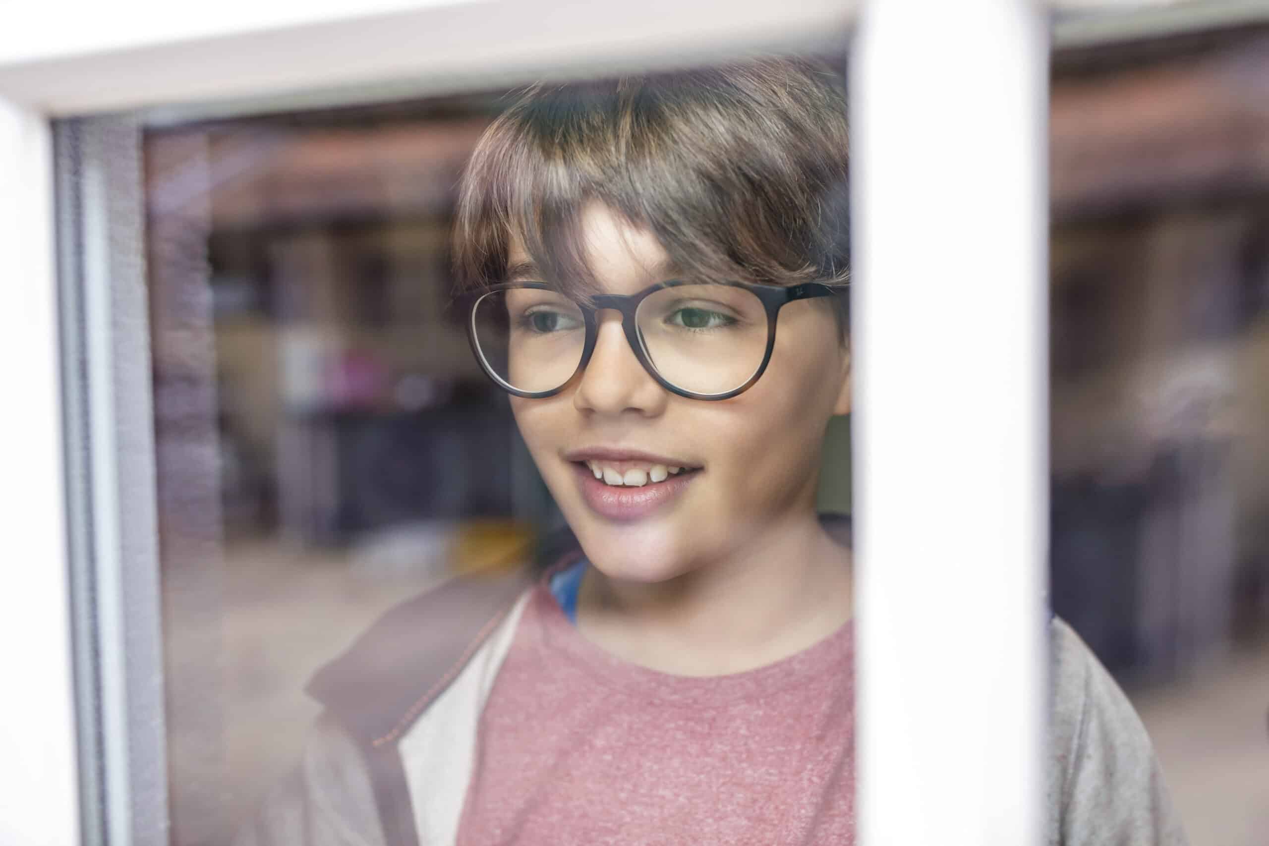 Ein Junge mit braunen Haaren und Brille lächelt, während er durch ein Fenster schaut. Er trägt ein rotes Hemd und eine helle Jacke. Das Glas des Fensters verwischt leicht den Blick auf den Hintergrund. © Fotografie Tomas Rodriguez