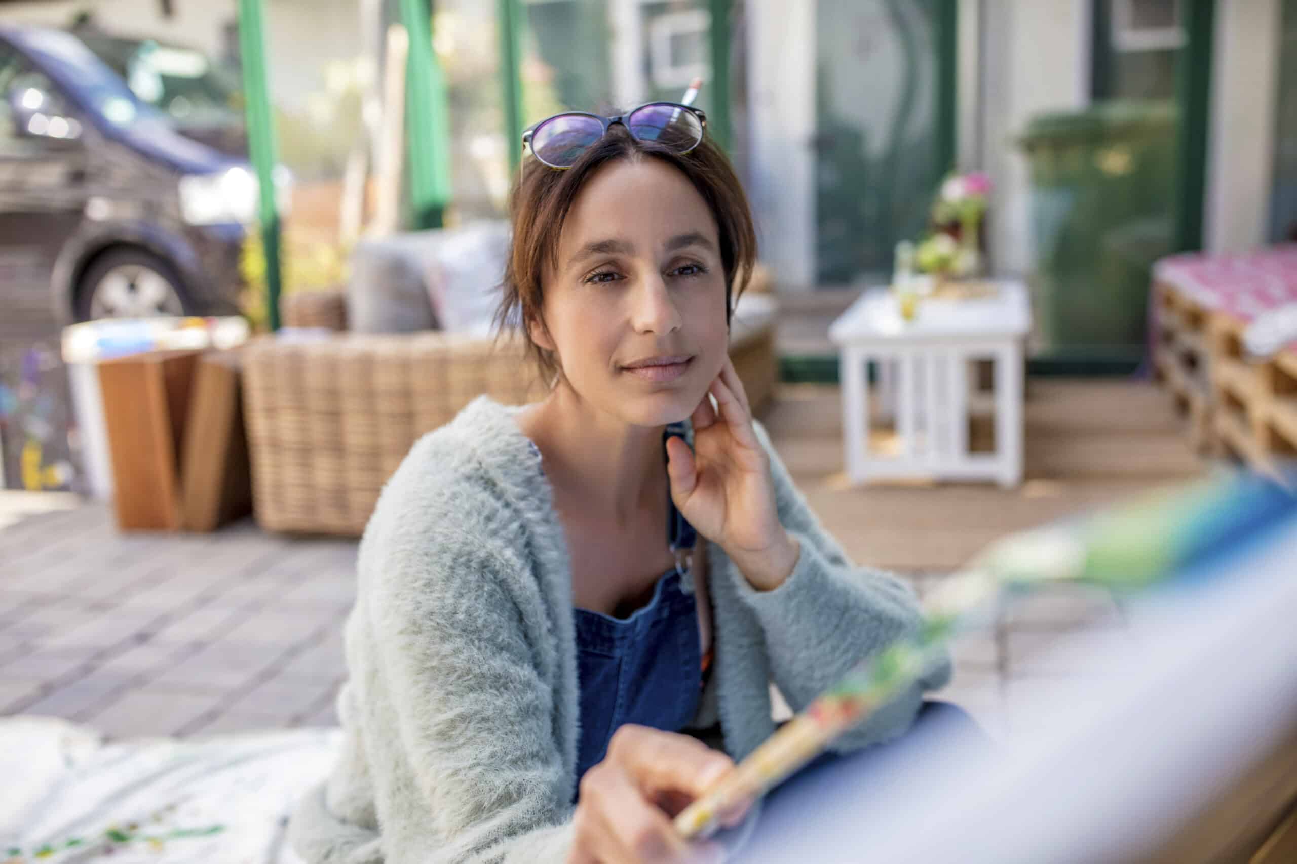 Eine Frau sitzt im Freien, trägt eine grüne Strickjacke und eine Brille auf dem Kopf. Sie malt auf einer Leinwand, im Hintergrund sind verschiedene Künstlerutensilien und Möbel zu sehen. Die Szene wirkt friedlich und kreativ. © Fotografie Tomas Rodriguez