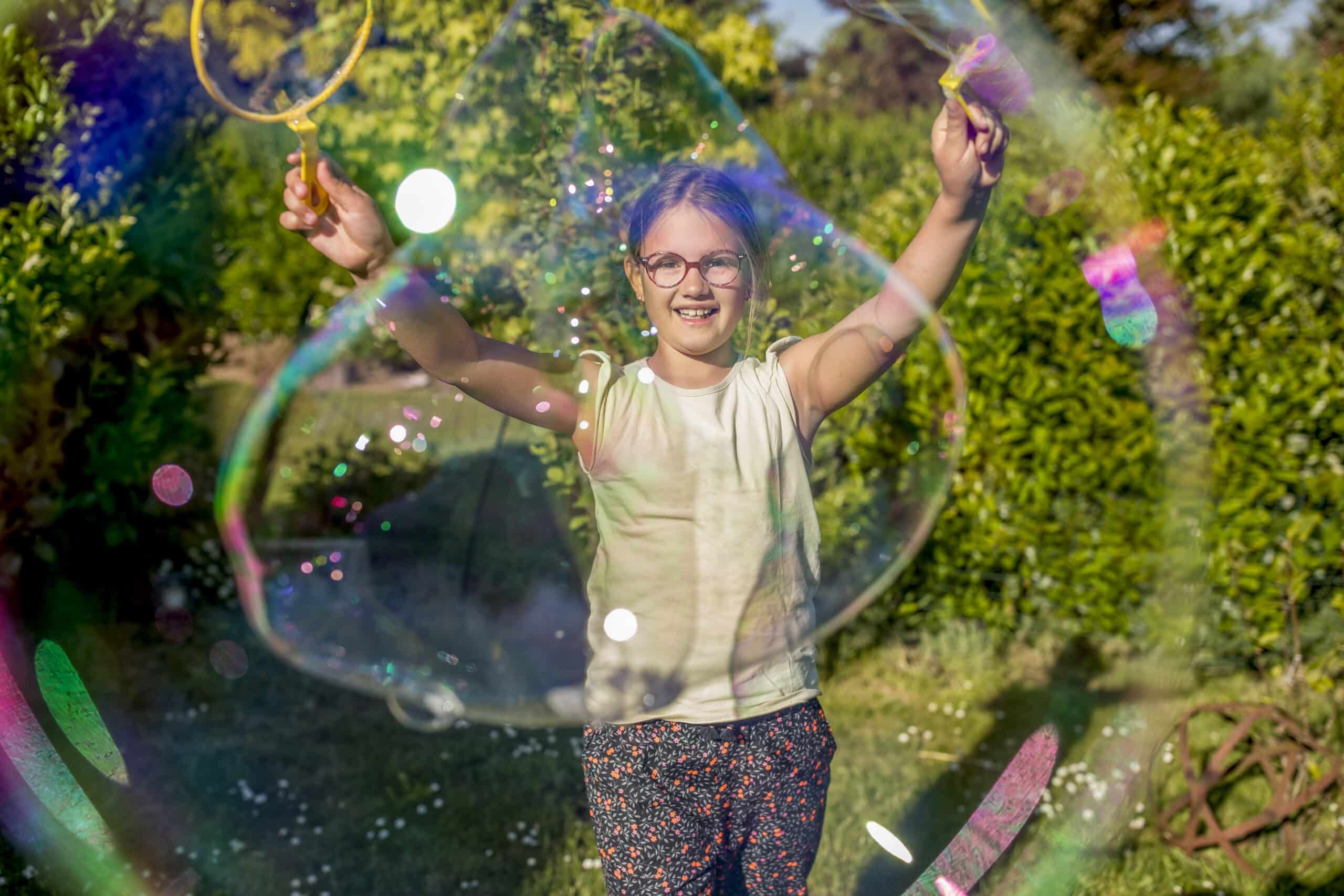 Ein kleines Kind mit Brille streckt freudig die Arme aus und hält an jedem Ende einen Seifenblasenstab. Es steht umgeben von einer riesigen Seifenblase vor einer üppigen, grünen Gartenkulisse. Die Sonne scheint hell und sorgt für eine fröhliche Atmosphäre. © Fotografie Tomas Rodriguez