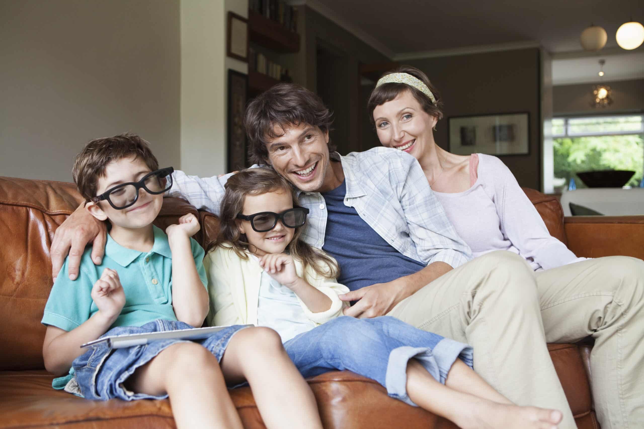 Eine vierköpfige Familie sitzt auf einem braunen Ledersofa, alle tragen 3D-Brillen und lächeln. Der Vater hat seinen Arm um Mutter und Tochter gelegt, während Sohn und Tochter, leger gekleidet, vorne sitzen. Im Hintergrund ist ein gemütliches, gut beleuchtetes Wohnzimmer zu sehen. © Fotografie Tomas Rodriguez
