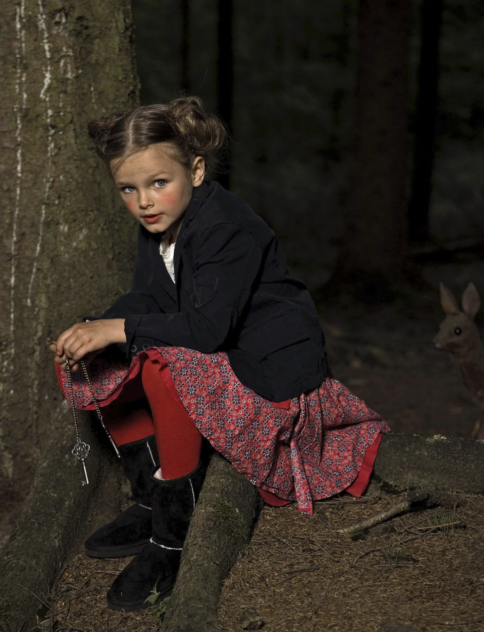 Ein junges Mädchen mit geflochtenen Haaren, einer dunklen Jacke, einem roten Rock und roten Leggings sitzt auf den Wurzeln eines Baumes in einem Wald. Sie hält einen Schlüsselbund mit einer Kette in der Hand. Im Hintergrund ist teilweise ein Hirsch zu sehen. © Fotografie Tomas Rodriguez