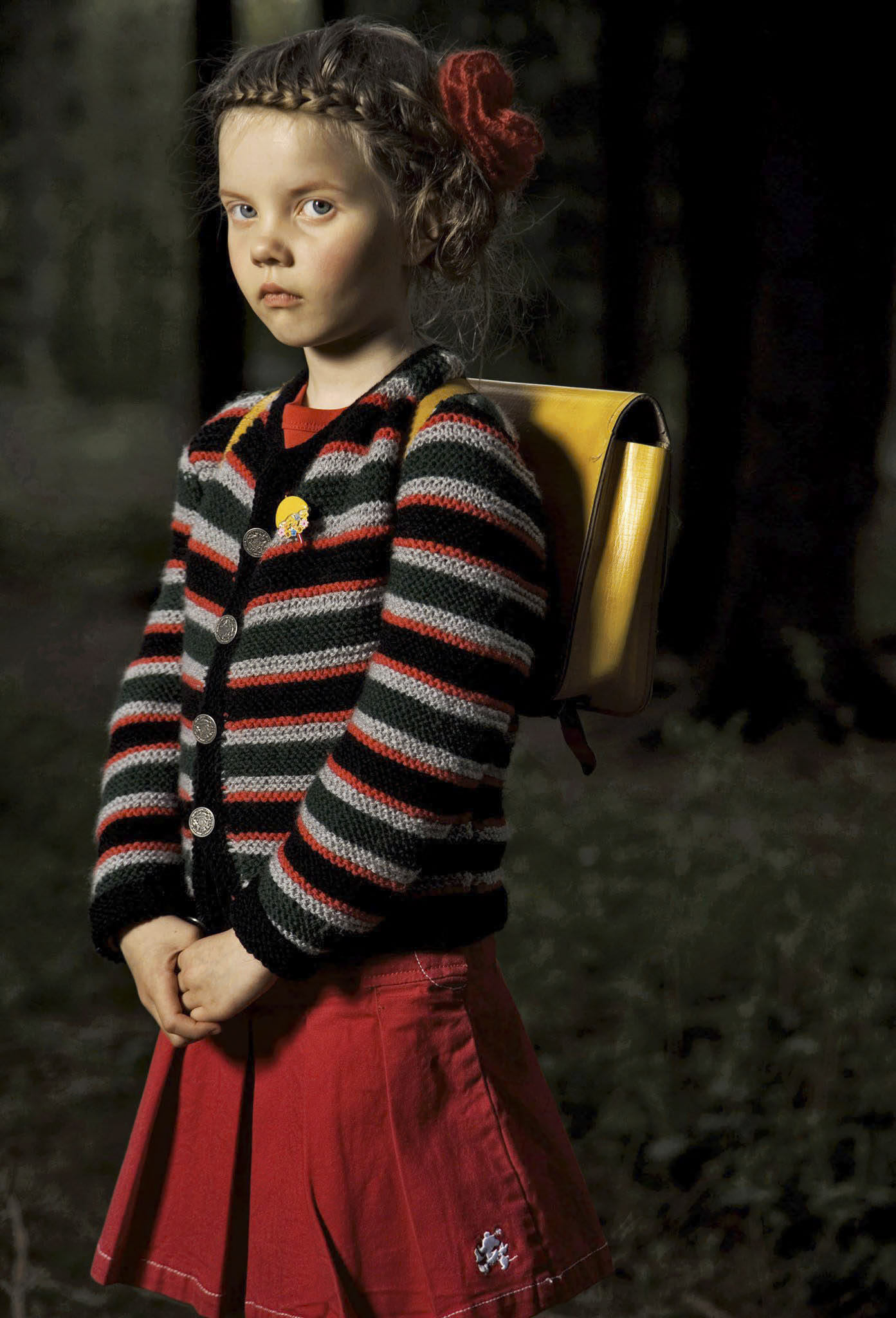 Ein junges Mädchen steht in einem schwach beleuchteten Wald. Sie trägt einen gestreiften Pullover und einen roten Rock. Sie trägt einen gelben Rucksack und hat ein rotes Haarband in ihrem frisierten Haar. Ihr Gesichtsausdruck ist ernst, während sie leicht zur Seite blickt. © Fotografie Tomas Rodriguez