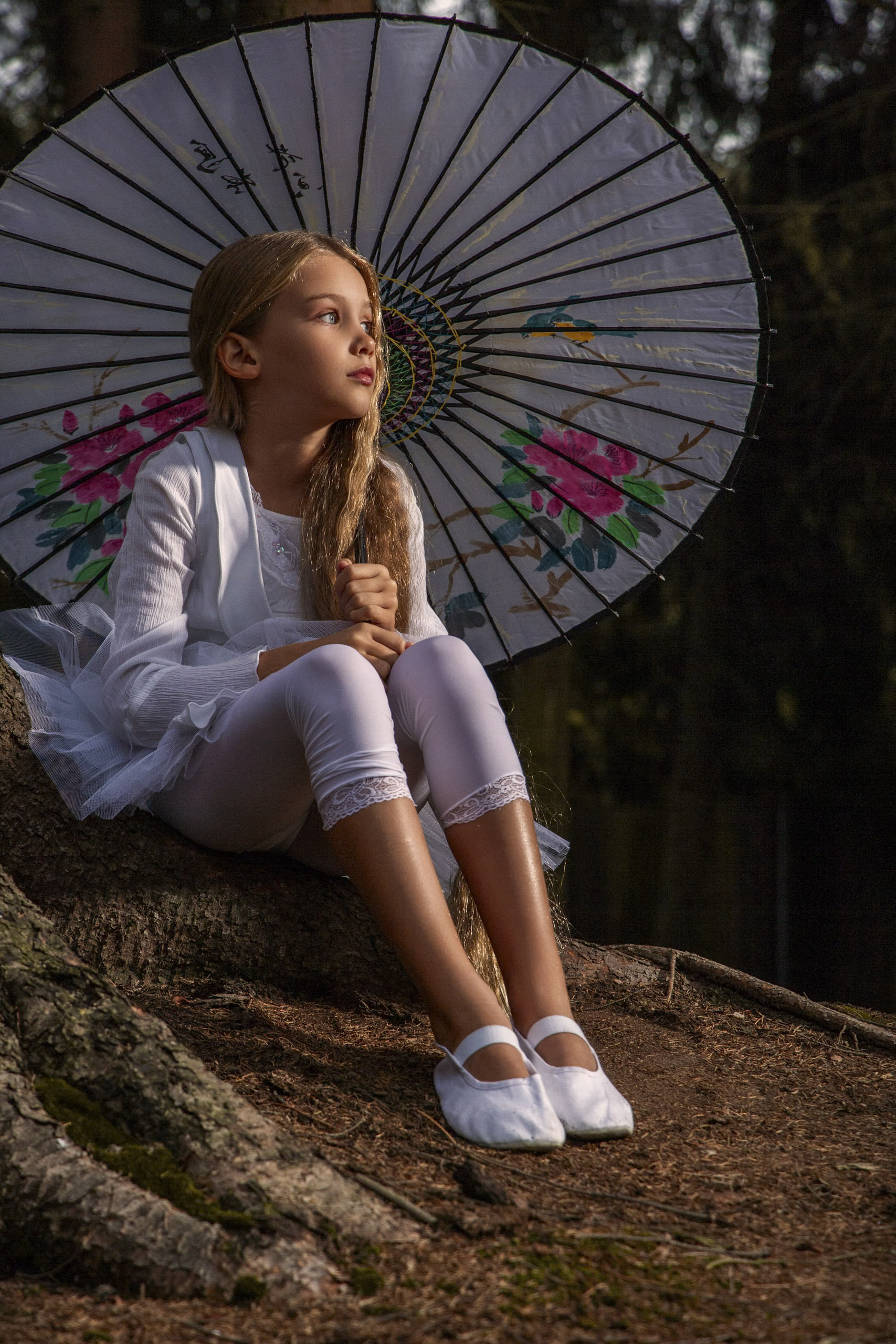 Ein junges Mädchen mit langen blonden Haaren und weißem Outfit sitzt auf einer Baumwurzel und hält einen dekorativen Regenschirm mit Blumenmustern in der Hand. Sie blickt nachdenklich in die Ferne. Im Hintergrund ist ein schwach beleuchteter Wald zu sehen. © Fotografie Tomas Rodriguez