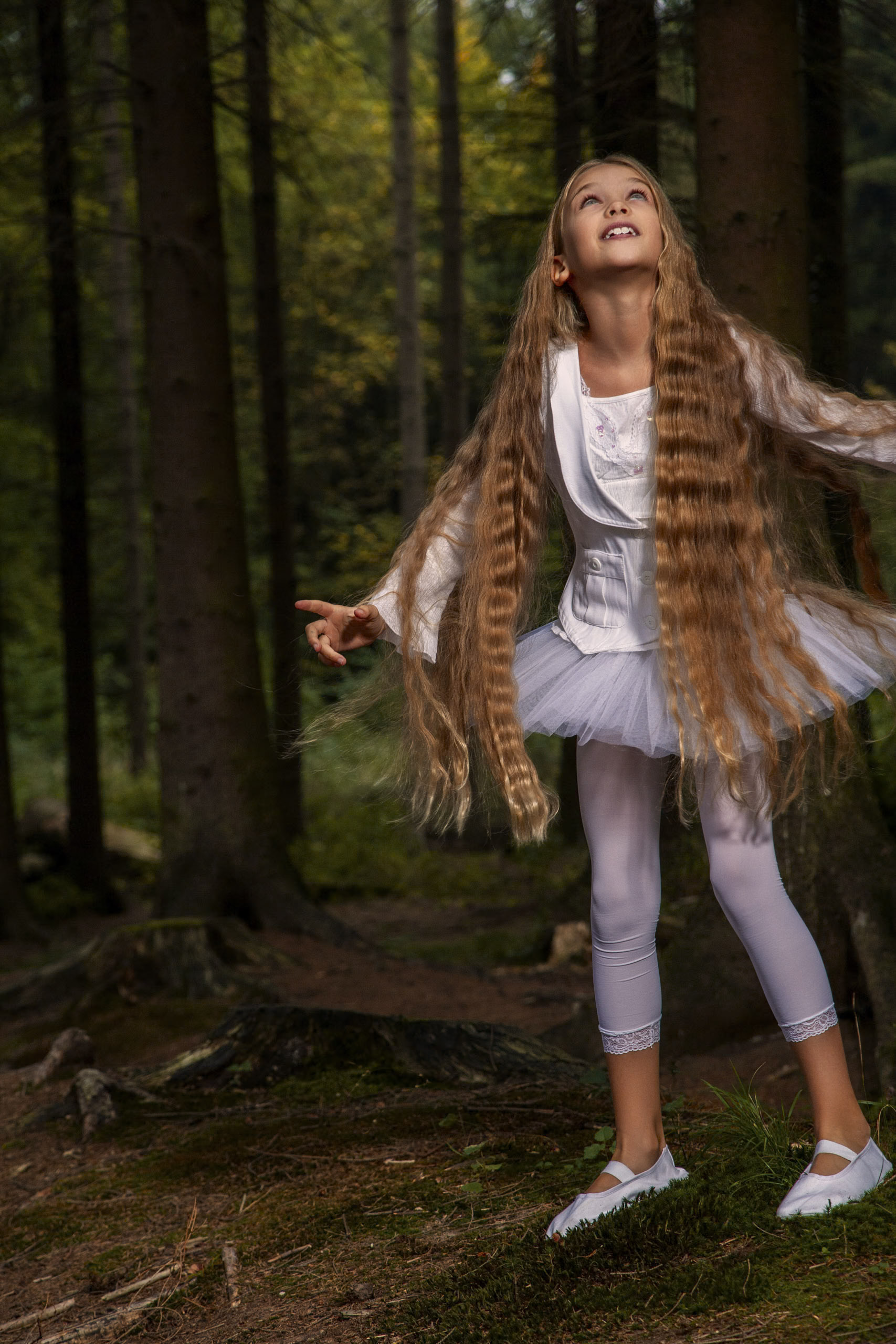 Ein junges Mädchen mit langen, welligen Haaren und einem weißen Ballettanzug steht in einem Wald. Sie blickt mit freudigem Gesichtsausdruck nach oben. Der Wald um sie herum ist üppig und dunkel, mit hohen Bäumen und einem grünen, moosbedeckten Boden. © Fotografie Tomas Rodriguez