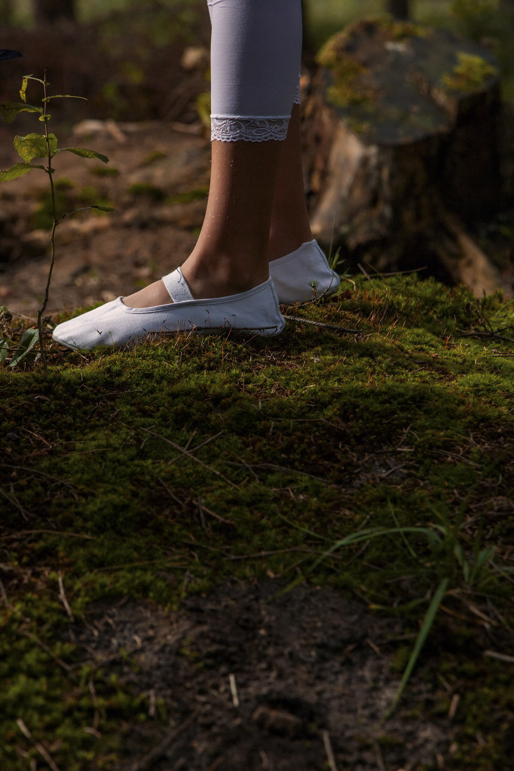 Eine Person in weißen Leggings mit Spitzenbesatz und weißen Ballerinas steht auf einem moosigen Waldboden, umgeben von kleinen Pflanzen und Baumstümpfen. © Fotografie Tomas Rodriguez