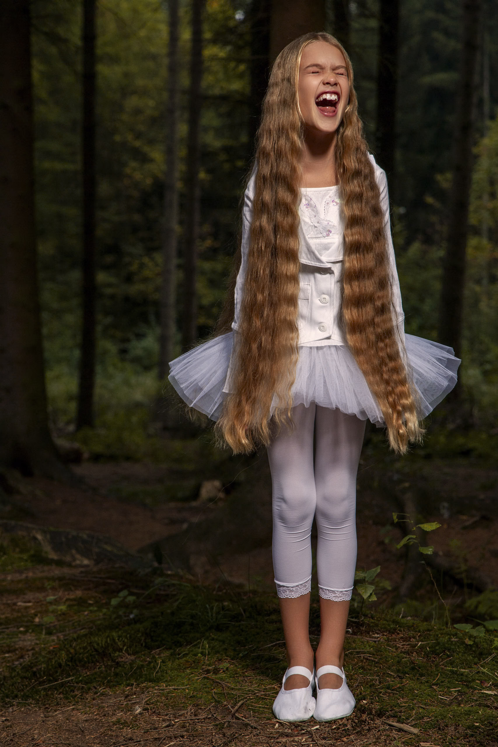 Ein junges Mädchen mit langen, welligen blonden Haaren steht in einem Wald und lacht fröhlich. Sie trägt ein weißes Ballett-Outfit, bestehend aus einem Tutu-Rock, einer Strickjacke und Leggings. Ihre Füße sind mit weißen Ballettschuhen geschmückt. © Fotografie Tomas Rodriguez