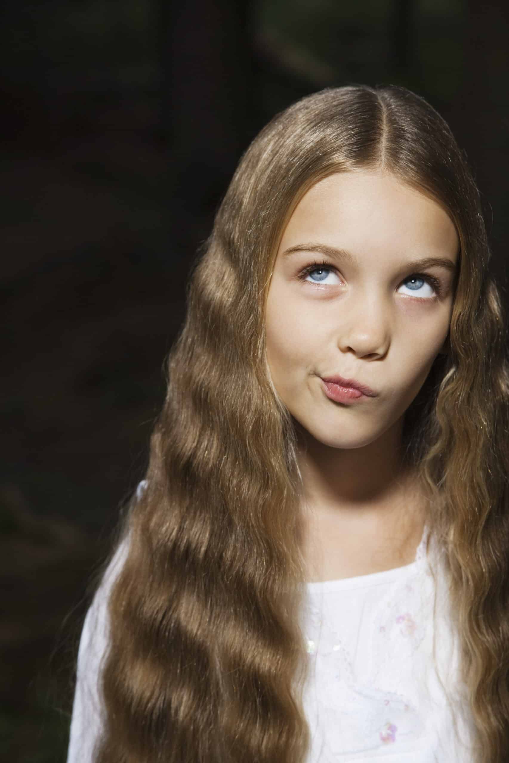 Ein junges Mädchen mit langen, welligen blonden Haaren und einem weißen Hemd blickt mit verspieltem Gesichtsausdruck nach oben, ihre Lippen sind leicht geschürzt, als würde sie tagträumen oder nachdenken. Der Hintergrund ist dunkel und verschwommen, wodurch sie hervorsticht. © Fotografie Tomas Rodriguez