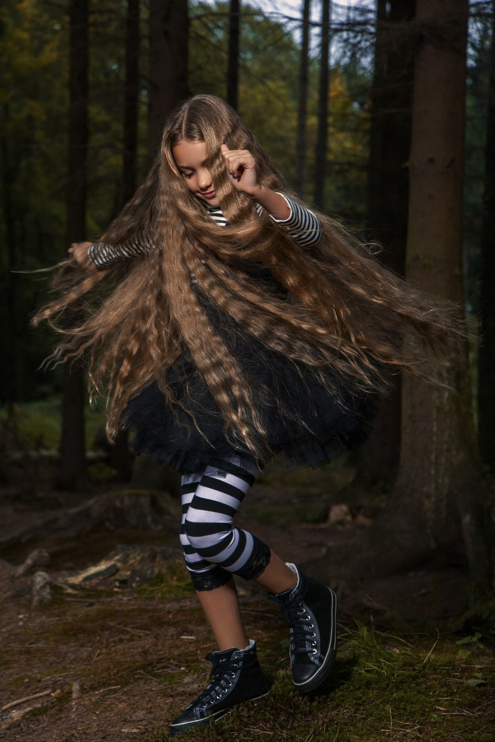 Ein junges Mädchen mit langen, welligen Haaren tanzt energisch durch einen Wald. Sie trägt ein gestreiftes Langarmshirt, einen schwarzen Rock, gestreifte Leggings und schwarze Turnschuhe. Im Hintergrund sind Bäume und Waldboden zu sehen. © Fotografie Tomas Rodriguez