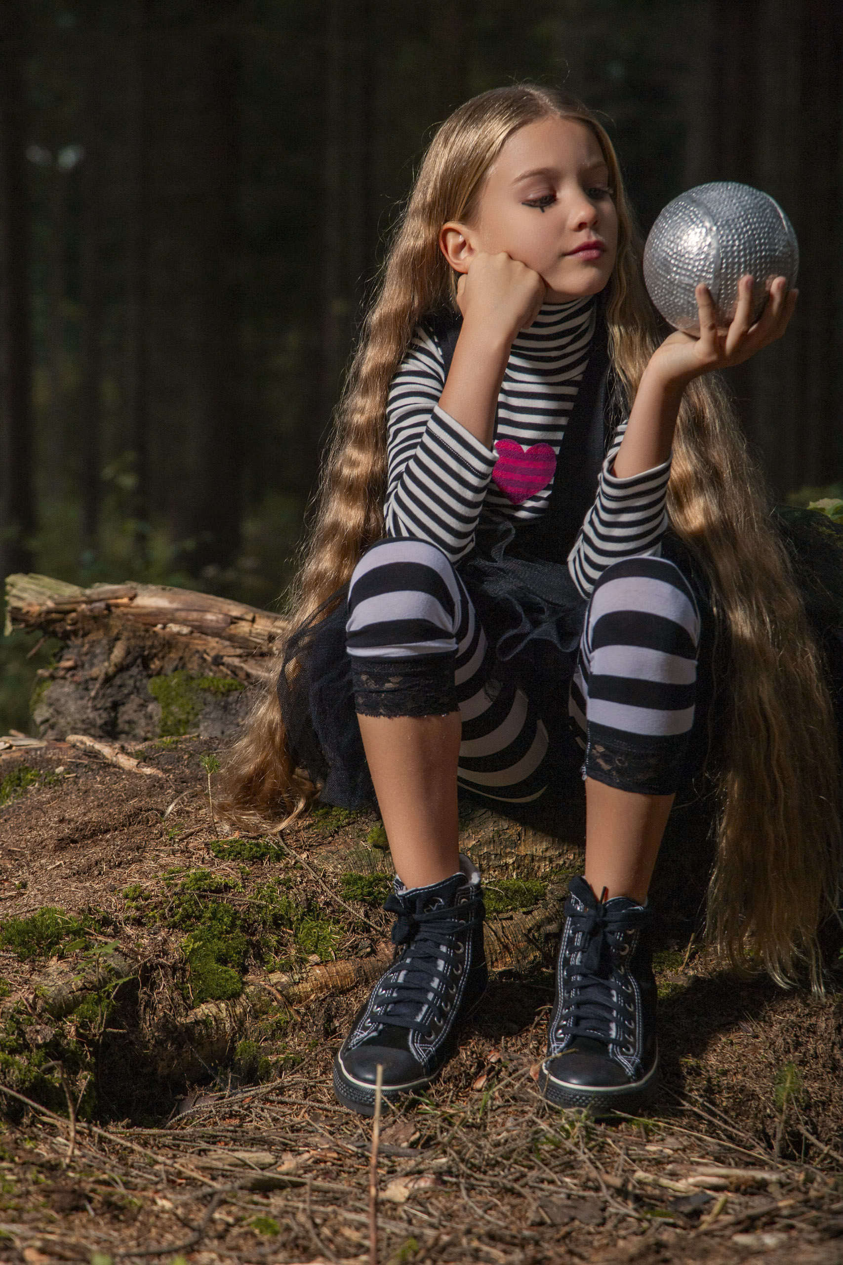 Ein junges Mädchen mit langen, welligen Haaren sitzt auf einem moosbedeckten Baumstumpf in einem Wald. Sie trägt schwarz-weiß gestreifte Kleidung und schwarze High-Top-Sneaker. Sie hält einen glänzenden silbernen Ball in der Hand, stützt ihren Kopf auf die andere Hand und betrachtet den Ball nachdenklich. © Fotografie Tomas Rodriguez