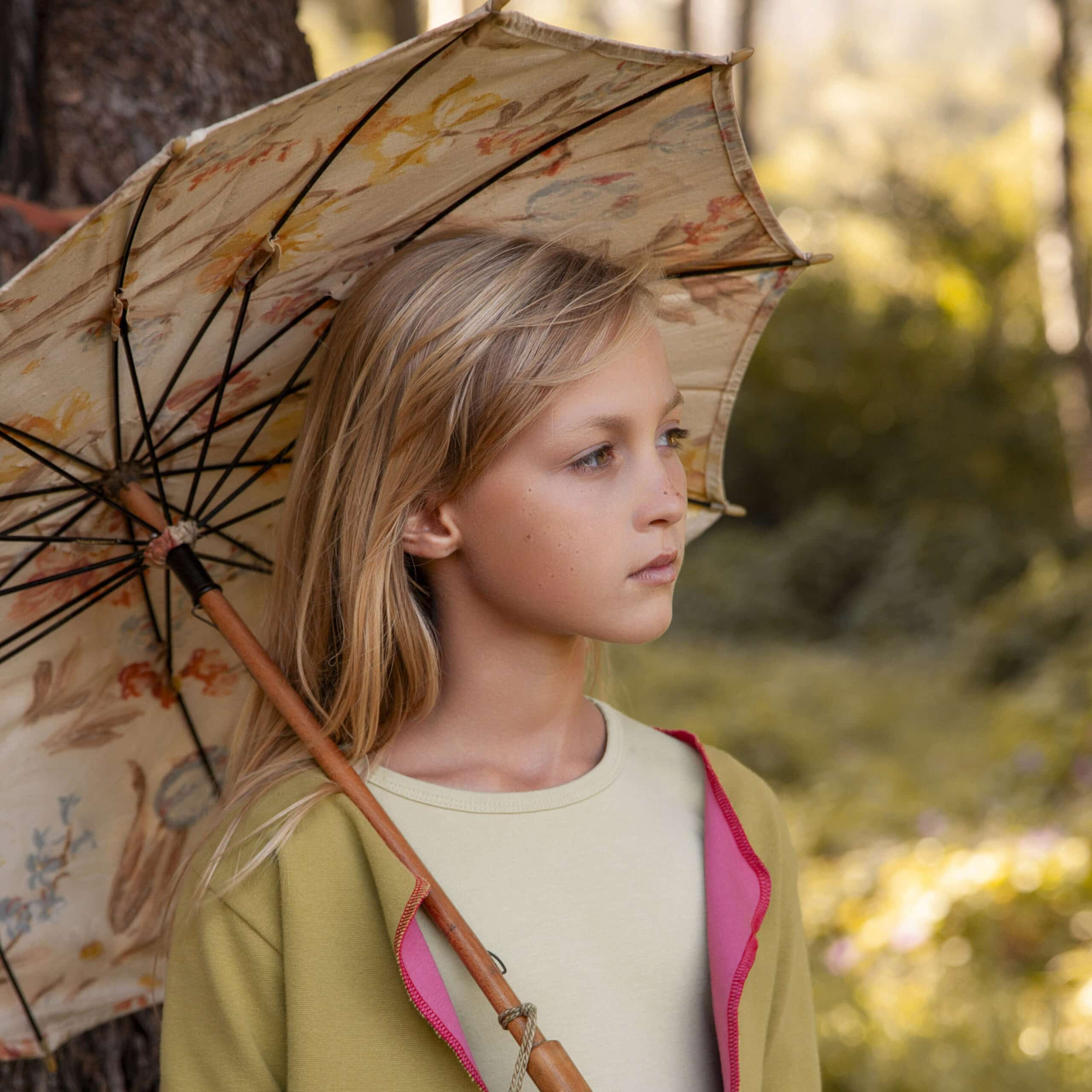 Ein junges Mädchen mit langen blonden Haaren steht in einem Wald und hält einen geblümten Sonnenschirm über der Schulter. Sie trägt eine grüne Jacke mit rosa Futter über einem hellen Hemd und blickt nachdenklich in die Ferne. Bäume und Grün umgeben sie. © Fotografie Tomas Rodriguez