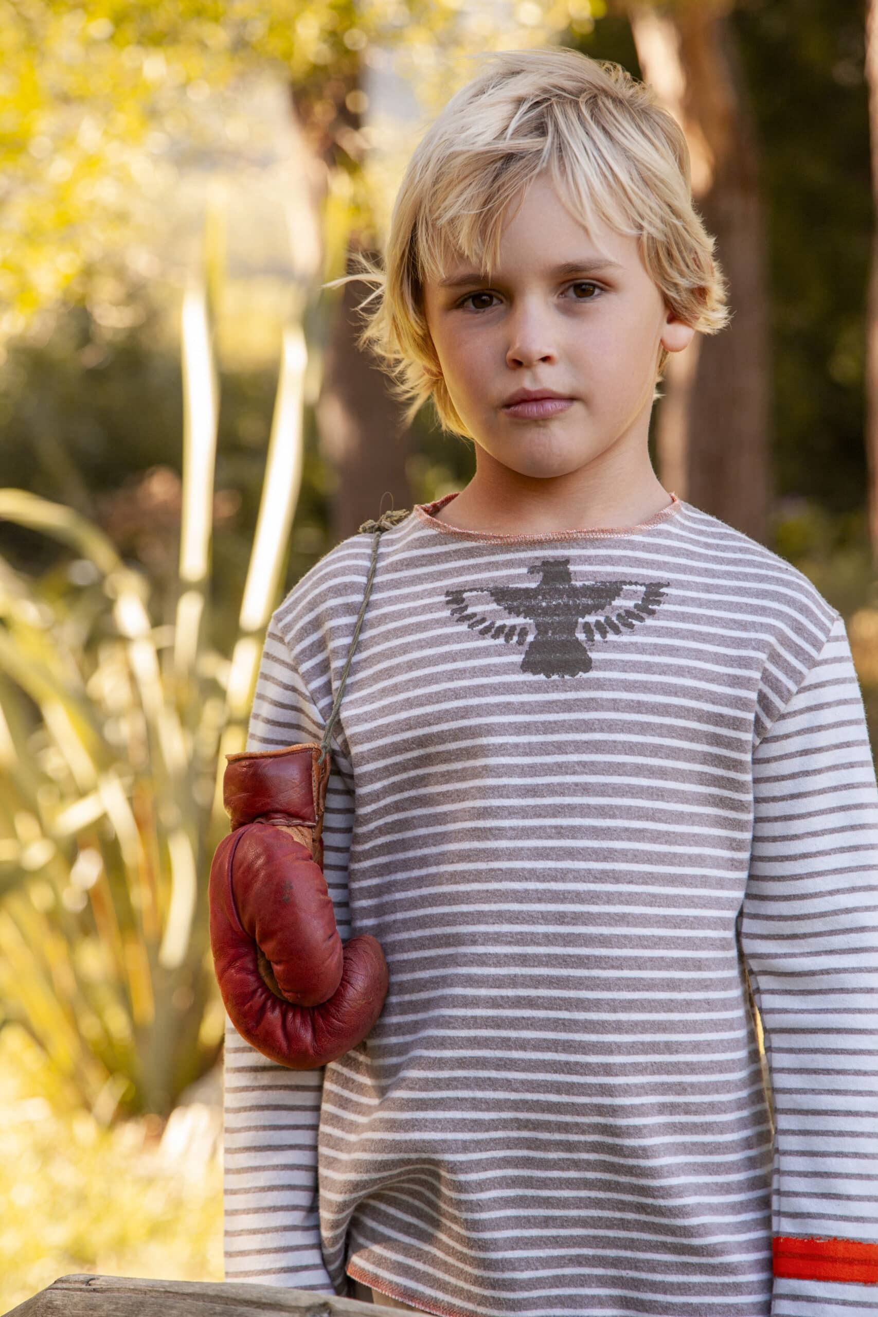 Ein kleines Kind mit blondem Haar steht im Freien und trägt ein grau-weiß gestreiftes Langarmshirt mit einem Adlermotiv. An seiner linken Schulter hängt ein roter Boxhandschuh. Im Hintergrund sind grüne Blätter und verschwommene Bäume zu sehen. © Fotografie Tomas Rodriguez