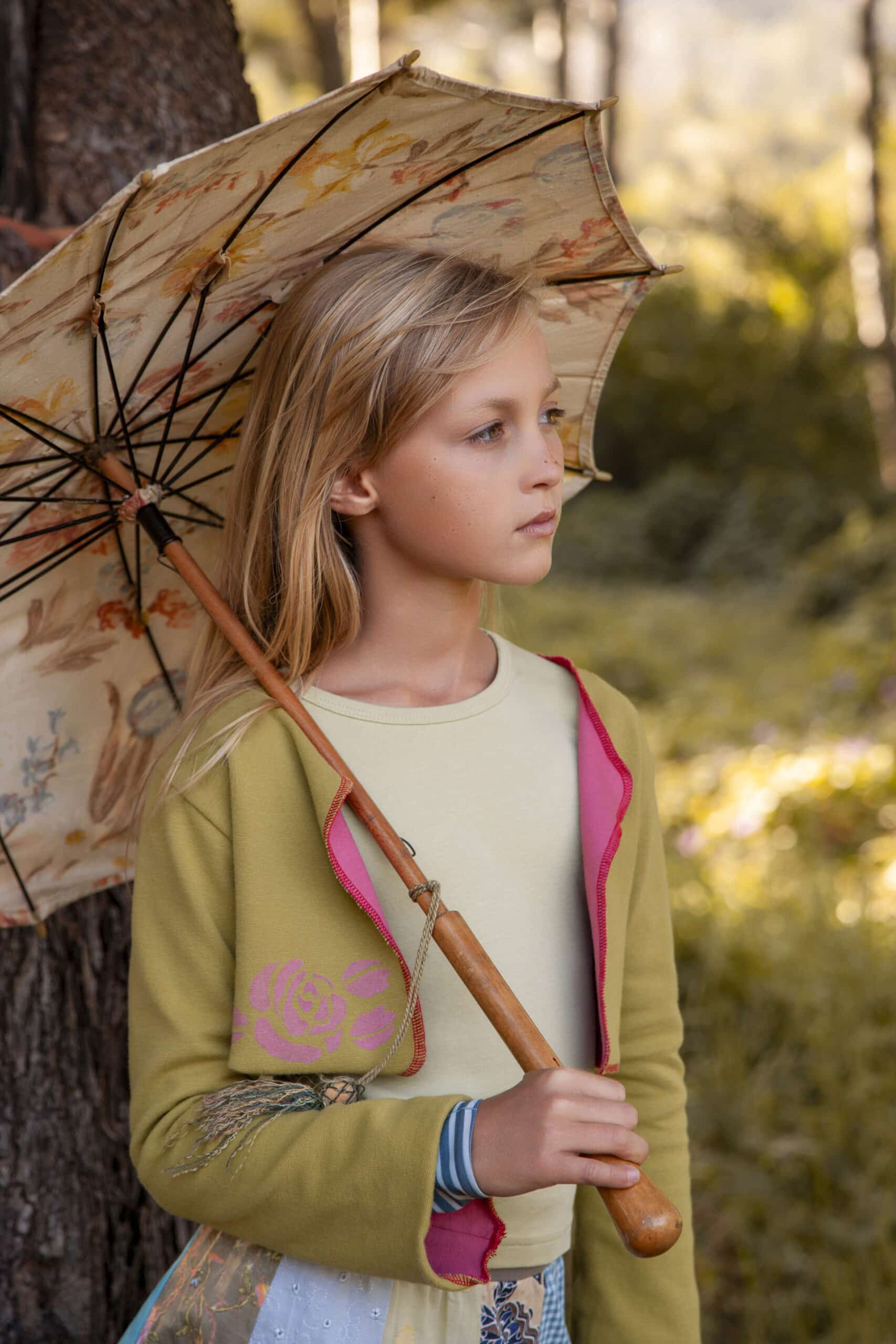 Ein Kind mit langen blonden Haaren steht im Freien und hält einen dekorativen Sonnenschirm über der Schulter. Das Kind trägt eine grüne Jacke mit rosa Futter und einem Blumendruck auf den Ärmeln. Es scheint nachdenklich in die Ferne zu blicken, mit Bäumen und Grün im Hintergrund. © Fotografie Tomas Rodriguez