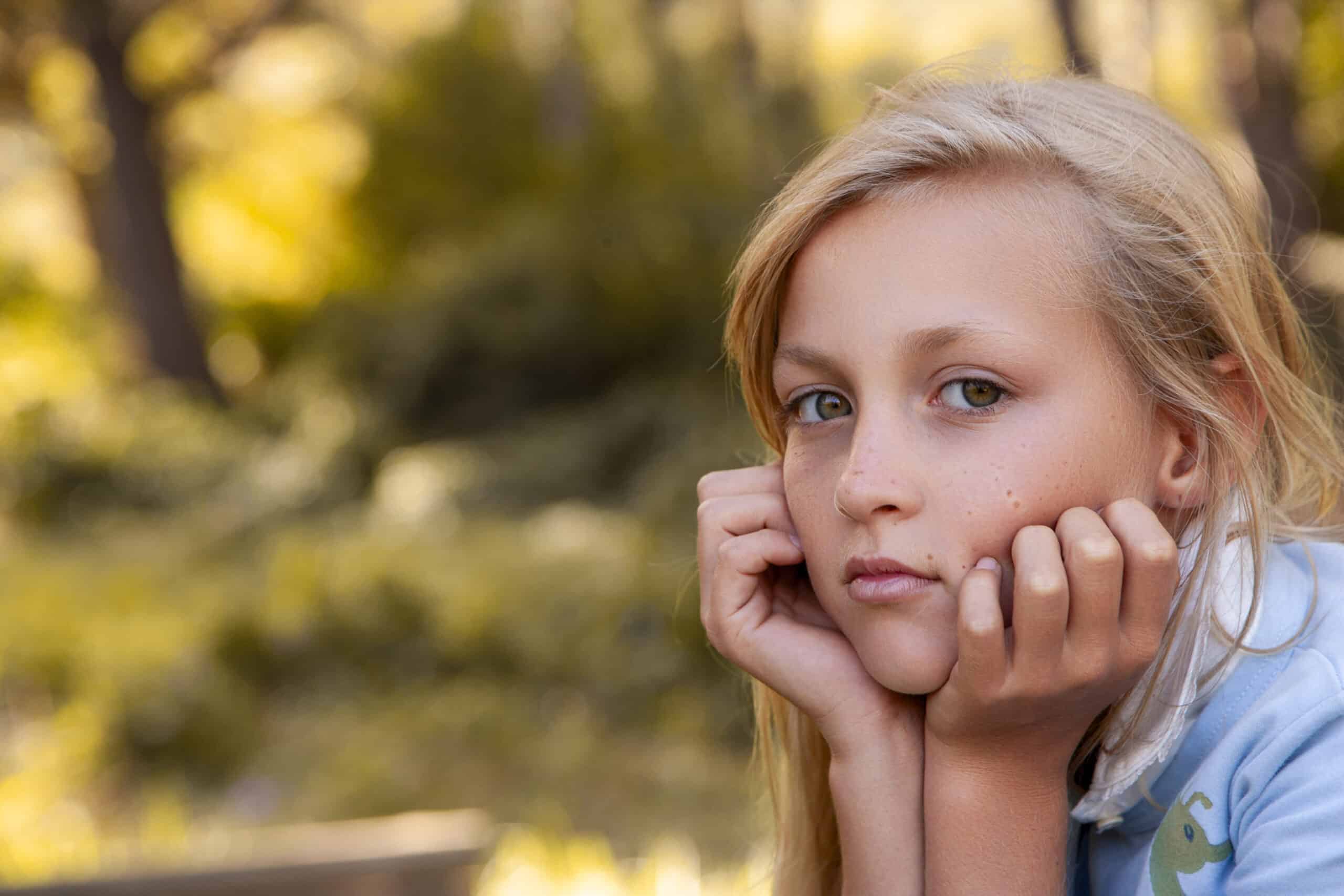 Ein junges Mädchen mit blondem Haar und nachdenklichem Gesichtsausdruck stützt ihr Kinn auf ihre Hände. Sie ist draußen vor einem verschwommenen Hintergrund aus Bäumen und Grün, was eine ruhige, natürliche Umgebung suggeriert. Sie trägt ein hellblaues Hemd und scheint in nachdenklicher Stimmung zu sein. © Fotografie Tomas Rodriguez