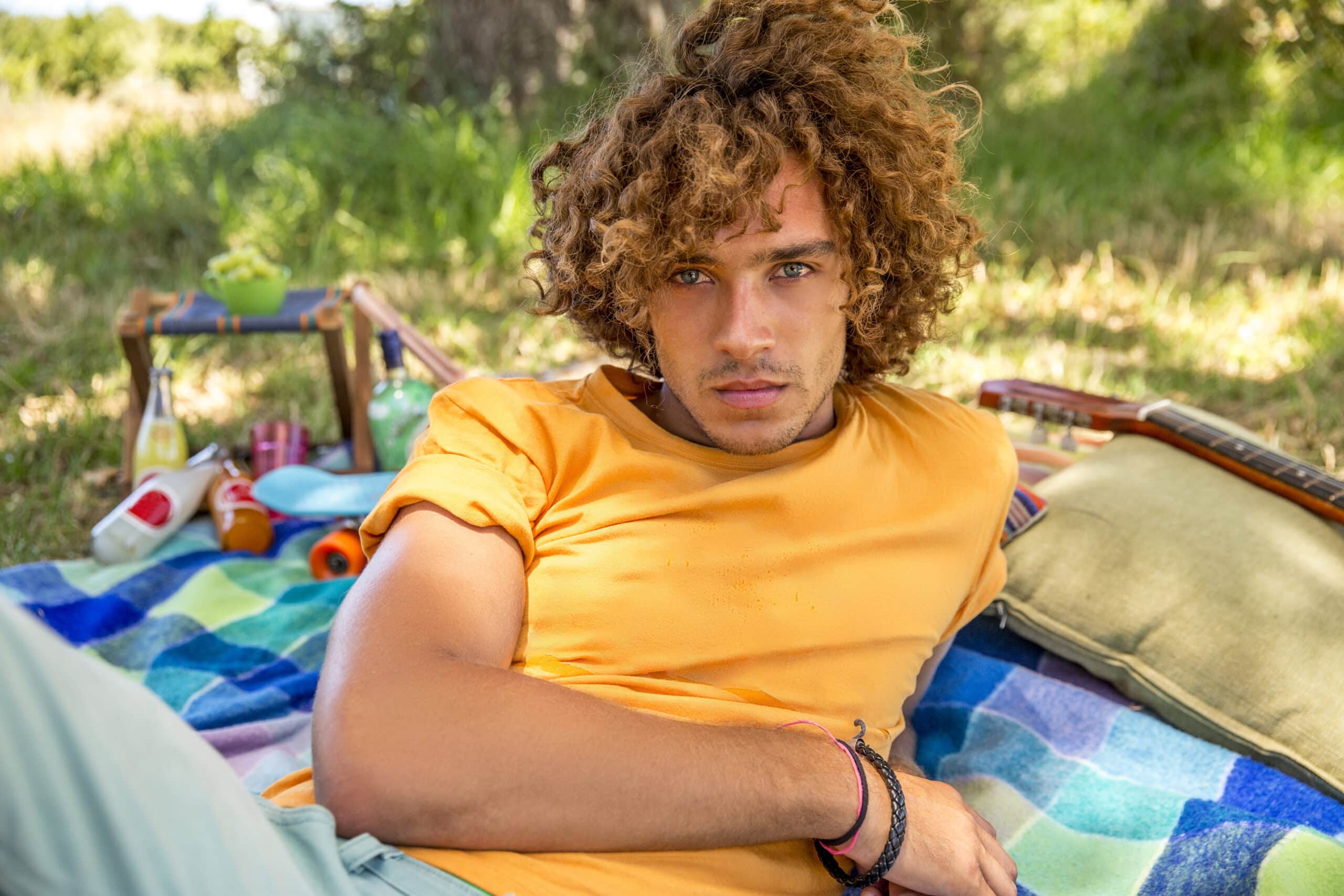 Ein junger Mann mit lockigem Haar und ernster Miene liegt auf einer bunten Decke in einem sonnigen Park, überall verstreut liegen Picknickutensilien. © Fotografie Tomas Rodriguez