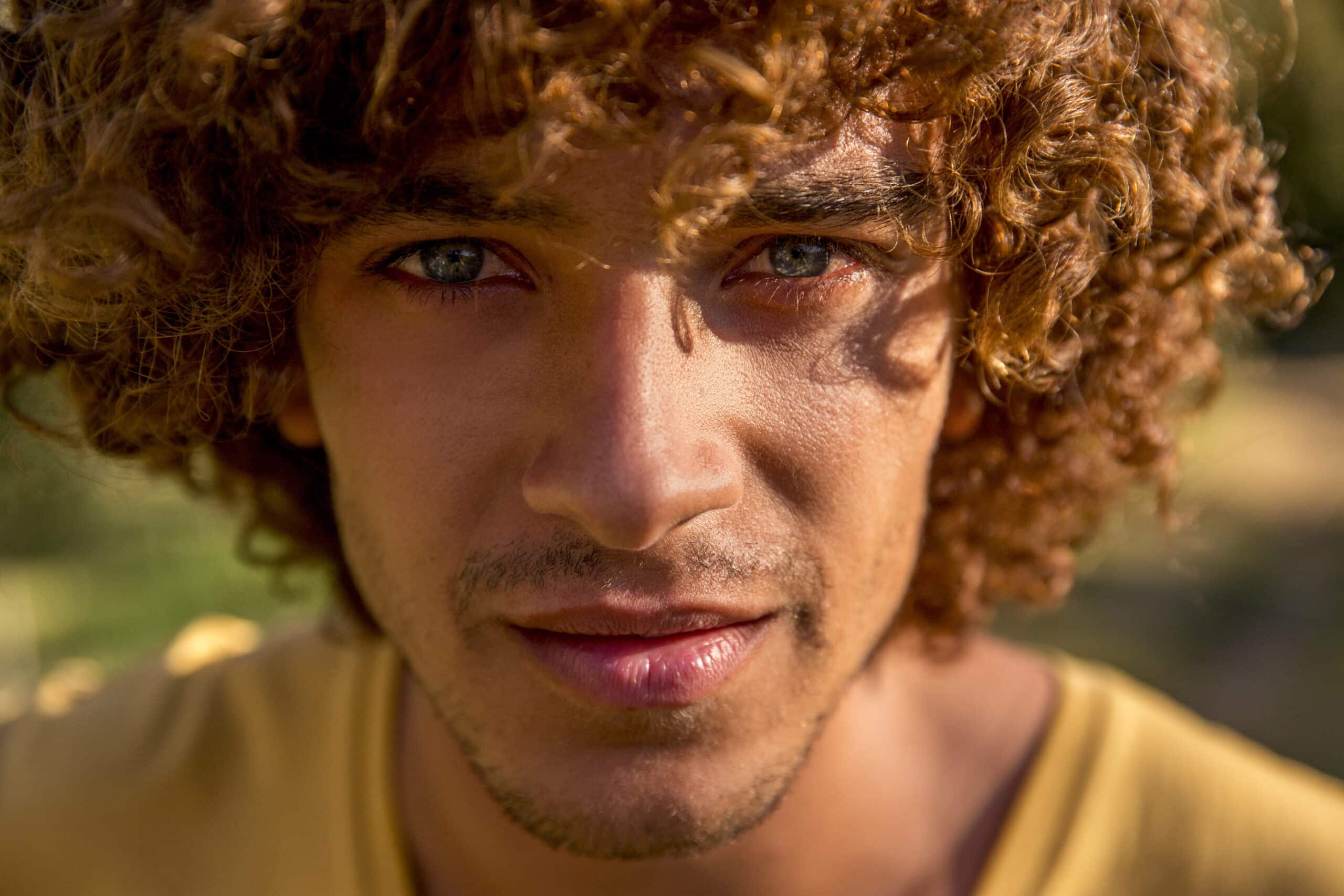 Nahaufnahme eines jungen Mannes mit lockigem Haar, wobei sein Gesicht im Sonnenlicht hervorgehoben wird. Er trägt ein gelbes Hemd und blickt direkt in die Kamera. © Fotografie Tomas Rodriguez