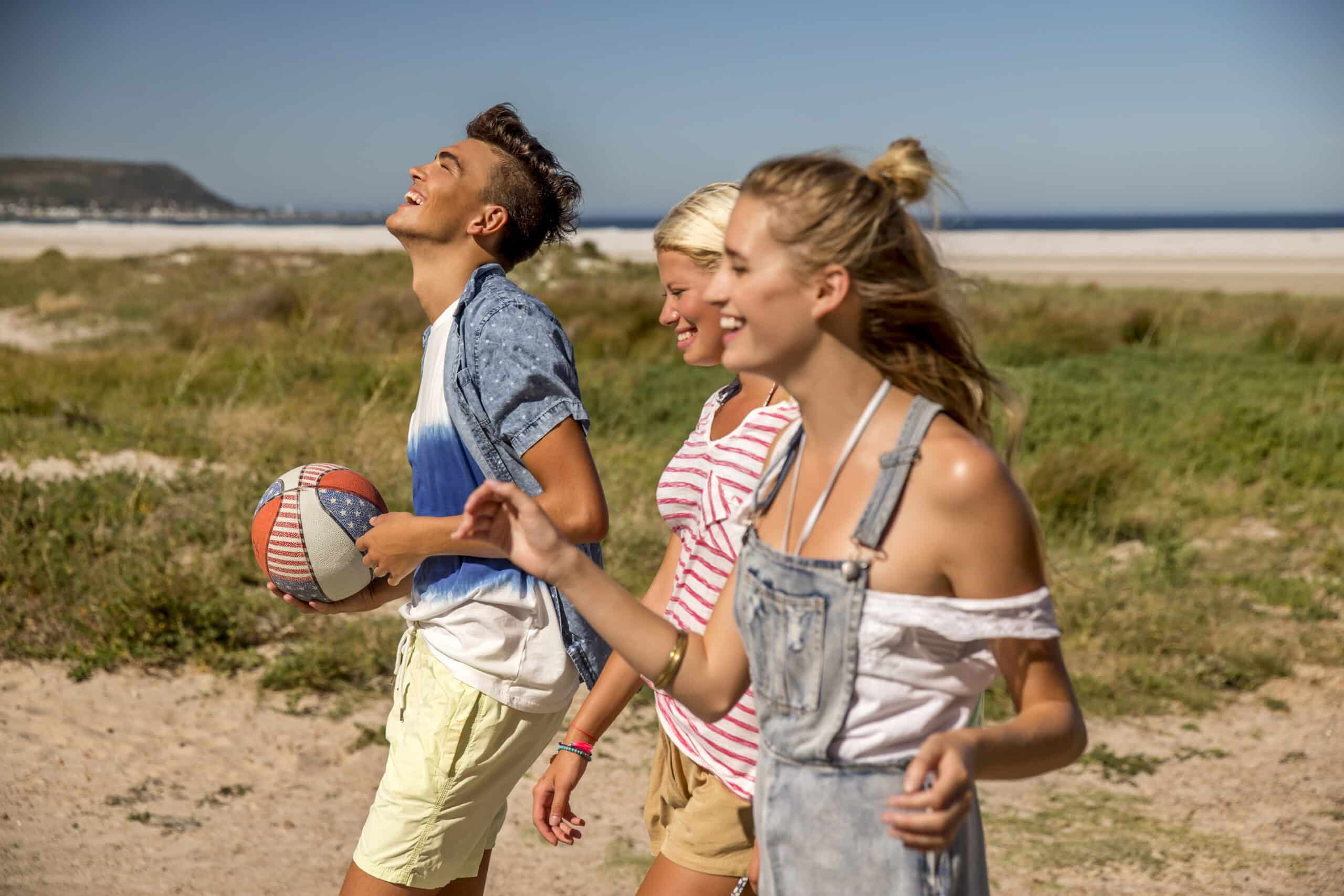 Drei junge Erwachsene lachen und gehen an einem Sandstrand spazieren, einer hält einen Volleyball. Sie genießen einen sonnigen Tag mit einem klaren blauen Himmel und fernen Hügeln im Hintergrund. © Fotografie Tomas Rodriguez