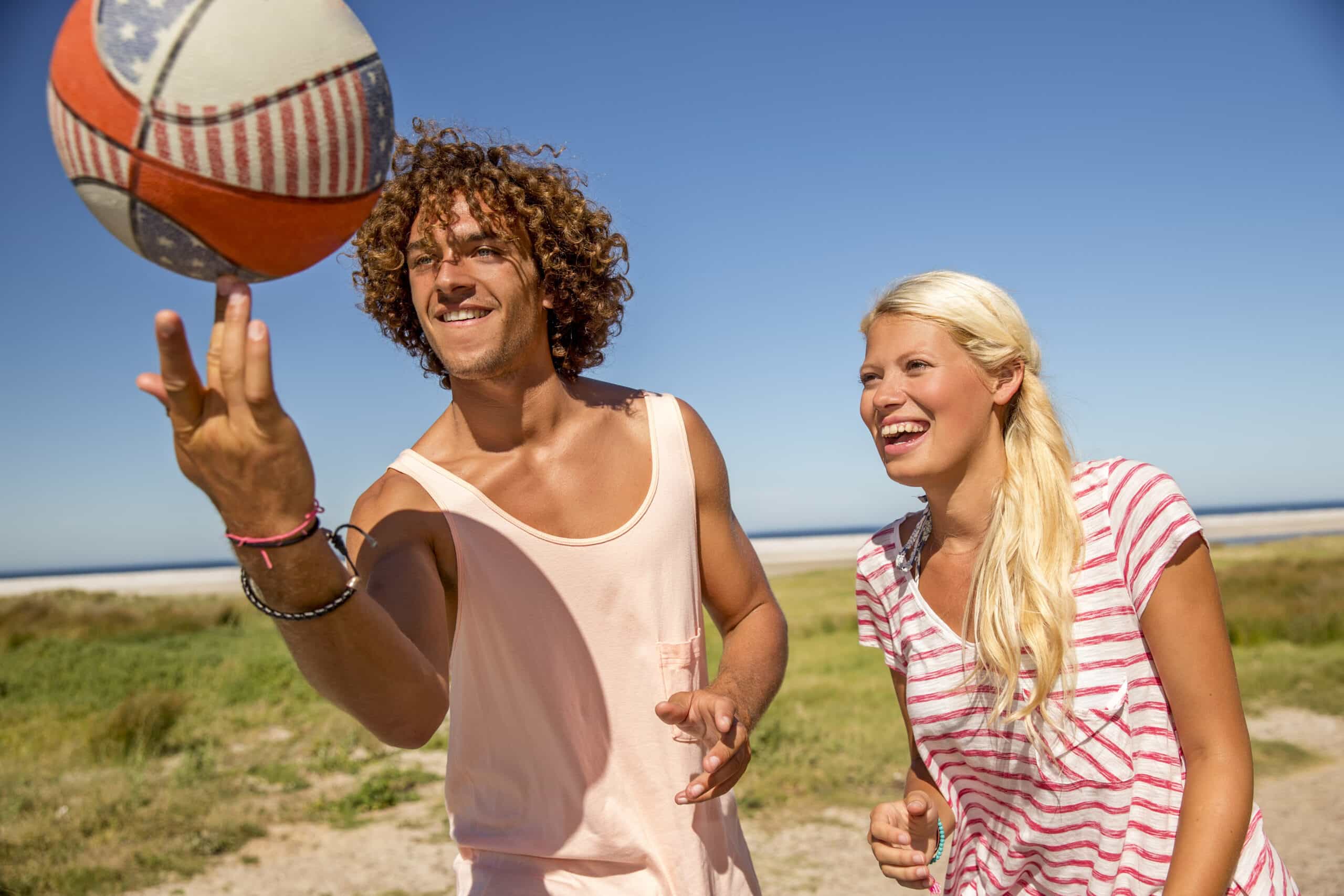 Ein junger Mann mit lockigem Haar, der einen Volleyball auf seinem Finger kreisen lässt, steht neben einer lächelnden blonden Frau in einem gestreiften Hemd an einem sonnigen Strand. © Fotografie Tomas Rodriguez