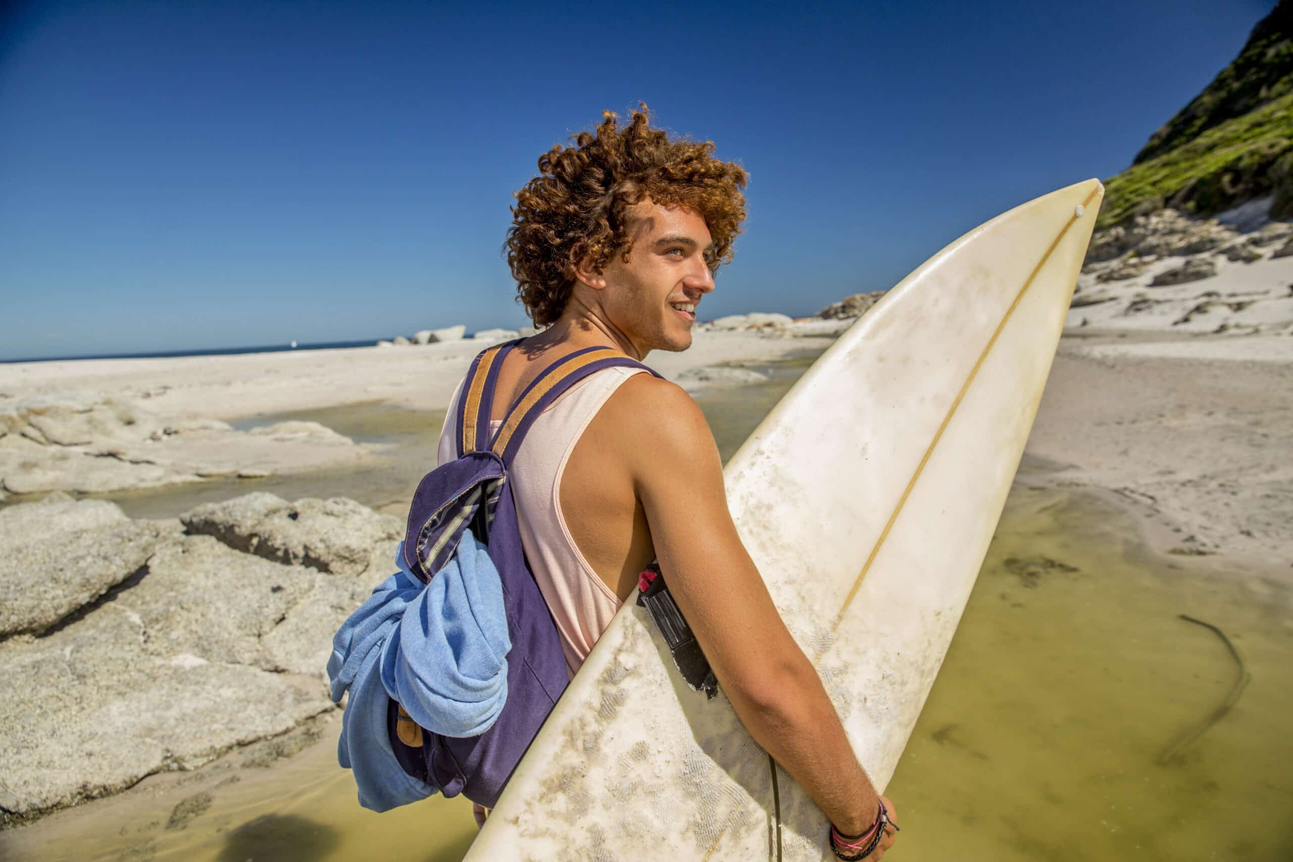 Ein junger Mann mit lockigem Haar, einem weißen Surfbrett und einem Rucksack auf dem Kopf, lächelt, während er an einem sonnigen Strand mit Felsen und dem Meer im Hintergrund spazieren geht. © Fotografie Tomas Rodriguez