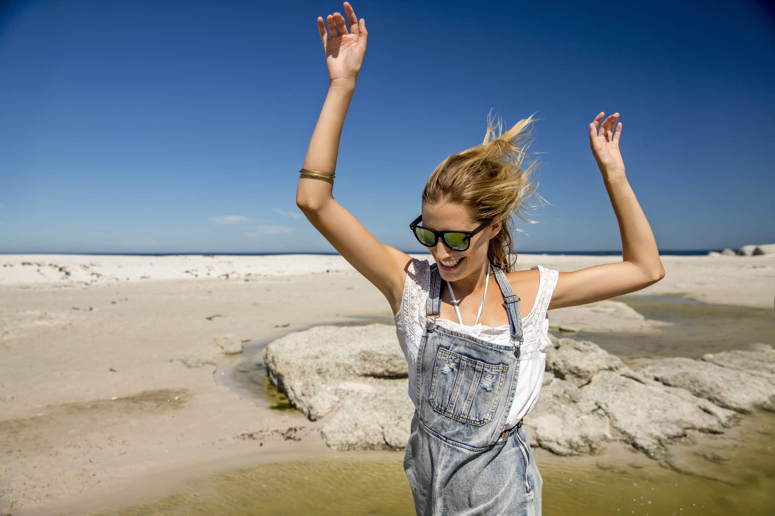 Eine fröhliche Frau in Jeans-Overall und Sonnenbrille streckt an einem sonnigen Strand mit weißem Sand und verstreuten Steinen jubelnd die Arme in die Höhe. © Fotografie Tomas Rodriguez