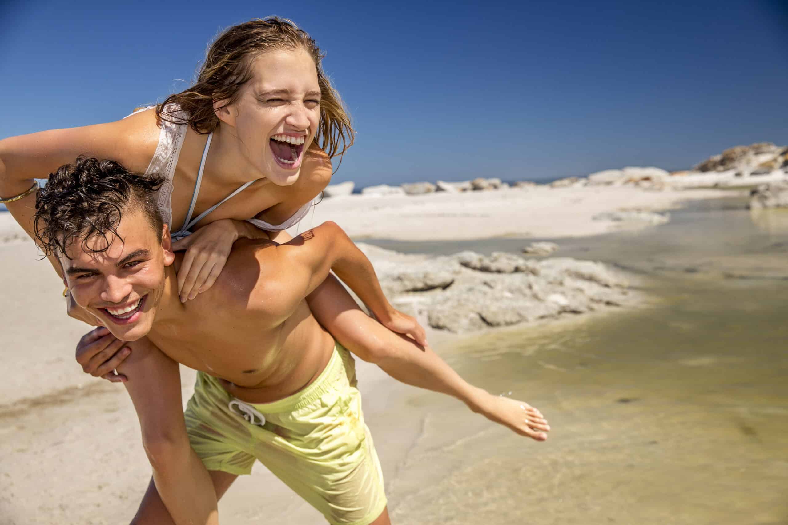 Eine fröhliche junge Frau reitet Huckepack auf einem lächelnden jungen Mann an einem sonnigen Strand. Beide tragen Badebekleidung und laufen verspielt am Ufer entlang. © Fotografie Tomas Rodriguez