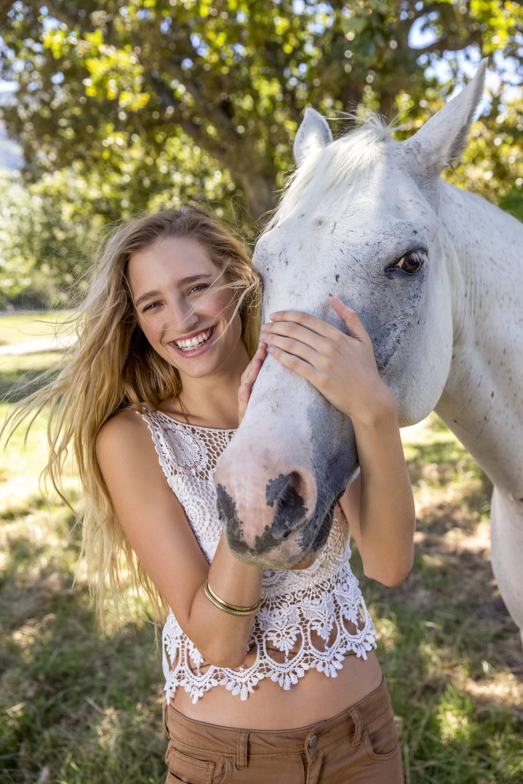 Eine fröhliche junge Frau mit langen blonden Haaren, einem Spitzentop und braunen Hosen bekleidet, umarmt verspielt ein weißes Pferd auf einer sonnigen Wiese. © Fotografie Tomas Rodriguez