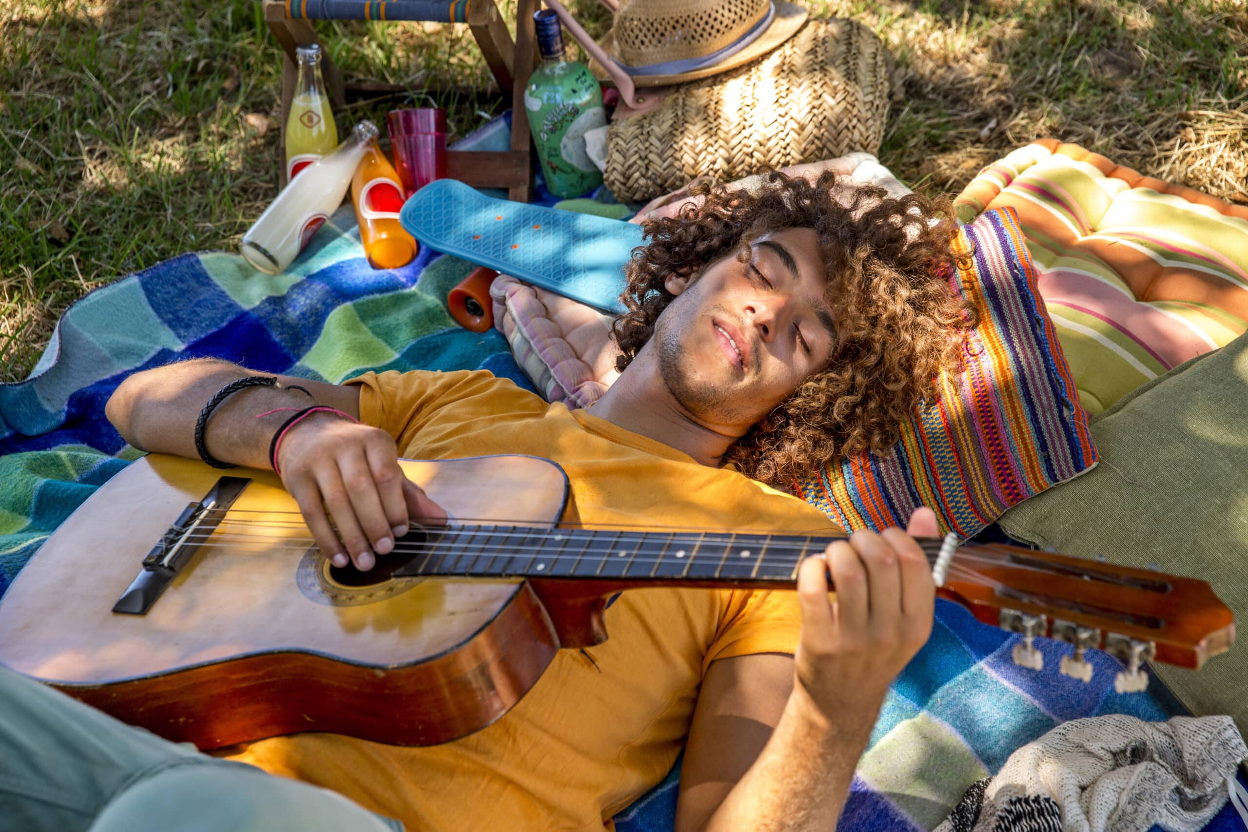 Ein junger Mann mit lockigem Haar liegt im Freien auf einer bunten Decke und hält eine Gitarre. Er hat die Augen geschlossen und genießt die Sonne, umgeben von einer gestrickten Tasche und anderen Picknicksachen. © Fotografie Tomas Rodriguez