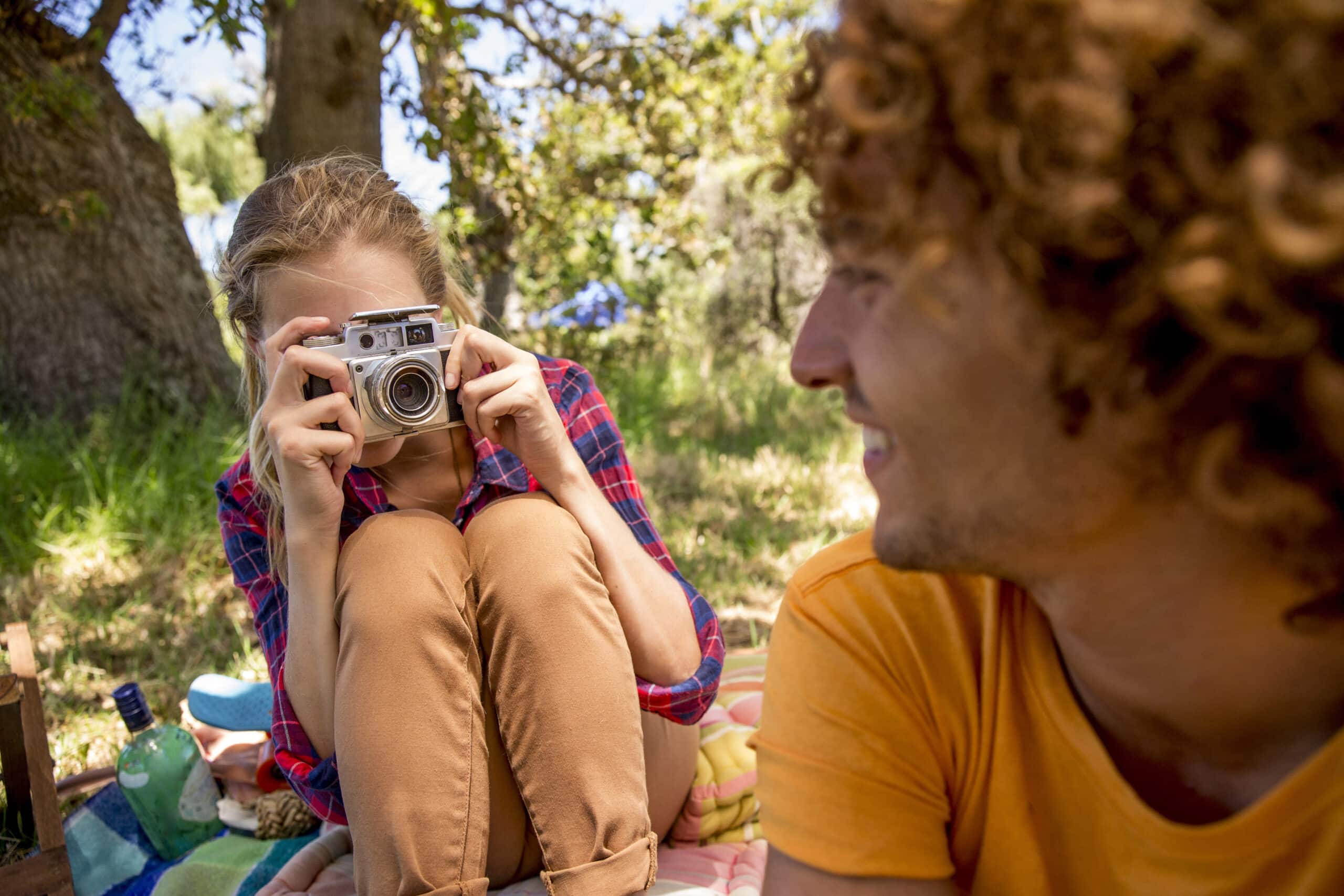 Eine Frau mit lockigem Haar fotografiert mit einer Vintage-Kamera einen lächelnden Mann mit lockigem Haar. Beide sitzen draußen bei einem Picknick unter schattigen Bäumen. © Fotografie Tomas Rodriguez