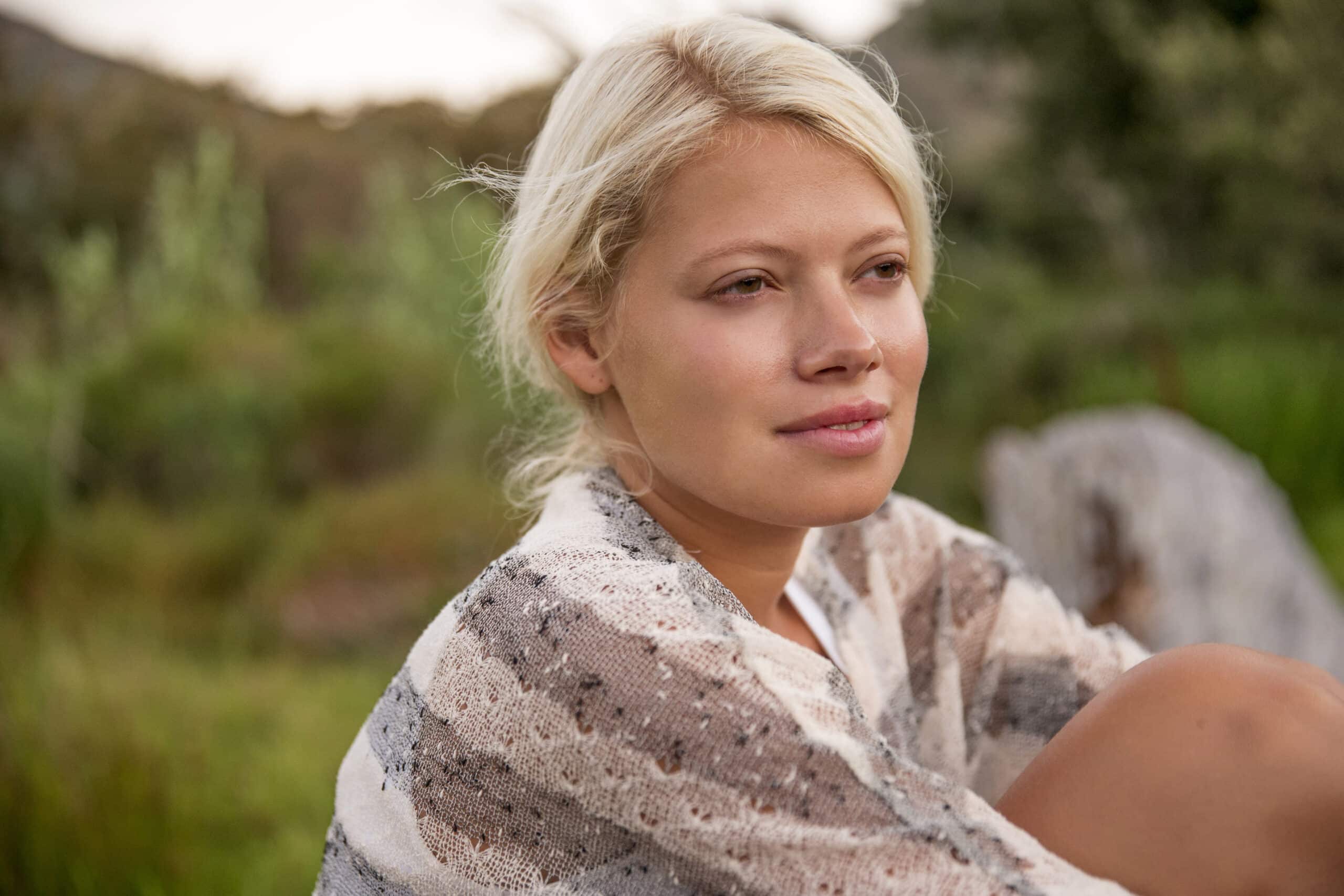 Eine heitere junge Frau mit blondem Haar sitzt mit nachdenklichem Gesichtsausdruck im Freien, in einen leichten, gemusterten Schal gehüllt, umgeben von üppigem Grün. © Fotografie Tomas Rodriguez