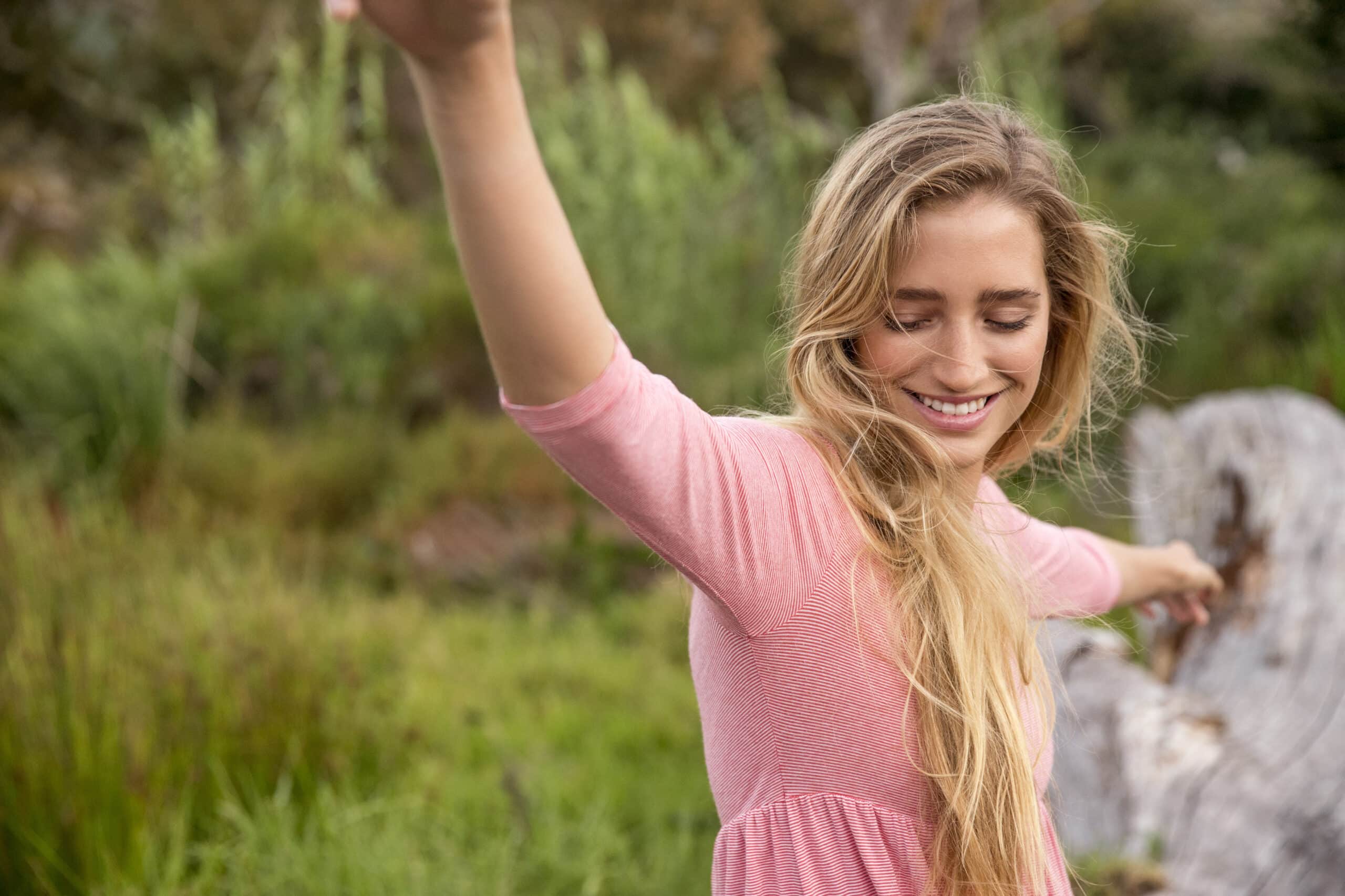 Eine fröhliche junge Frau mit langen blonden Haaren und einem rosa Oberteil streckt ihre Arme in einer natürlichen Umgebung mit viel Grün im Hintergrund aus. © Fotografie Tomas Rodriguez