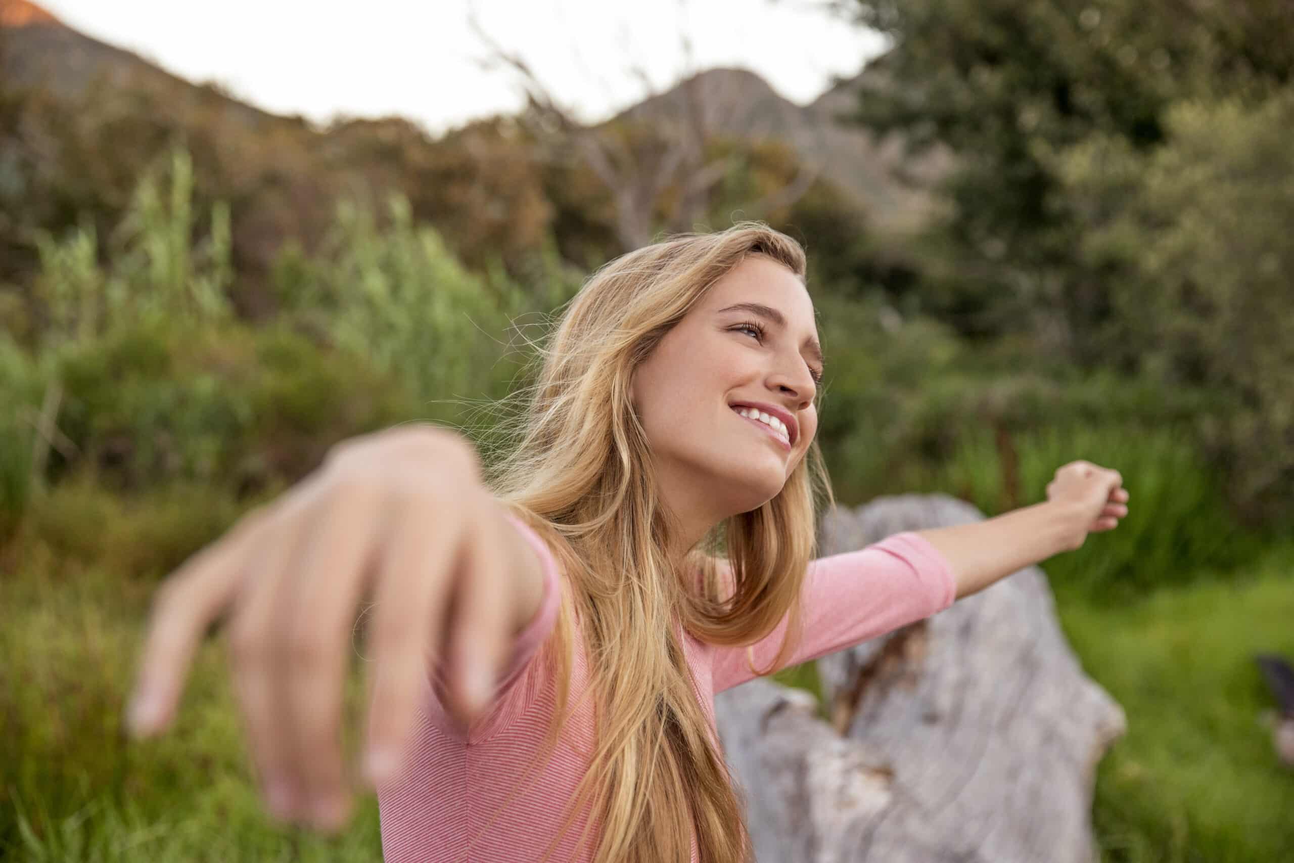 Eine fröhliche junge Frau mit langem blondem Haar zeigt in die Kamera und lächelt breit vor einer üppigen, grünen Außenkulisse mit Hügeln im Hintergrund. © Fotografie Tomas Rodriguez