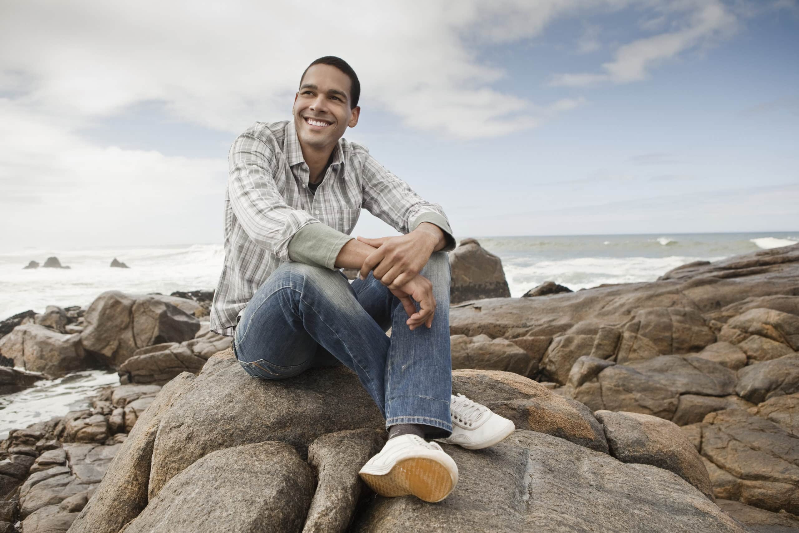 Ein lächelnder Mann sitzt in einem karierten Hemd und Jeans auf felsigem Küstengelände, im Hintergrund das Meer und ein bewölkter Himmel. © Fotografie Tomas Rodriguez