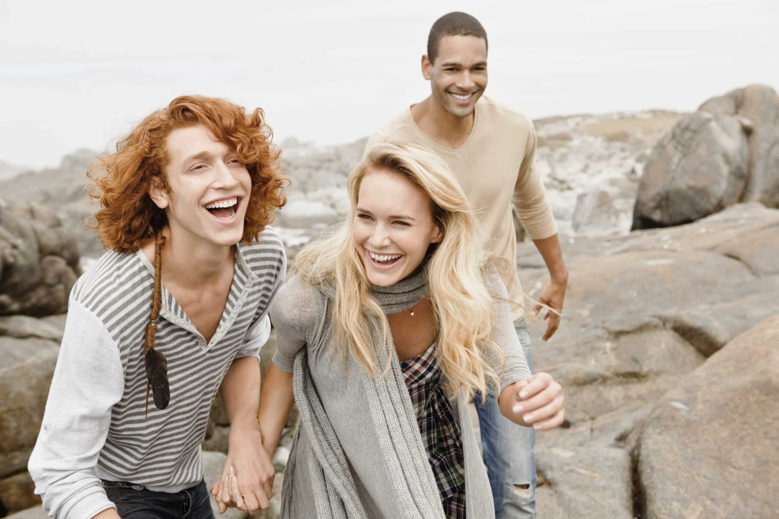 Drei Freunde lachen und laufen an einem steinigen Strand entlang, im Hintergrund ein bewölkter Himmel. Zwei Frauen und ein Mann sind in legere, bequeme Kleidung gekleidet und genießen einen lustigen Tag im Freien. © Fotografie Tomas Rodriguez