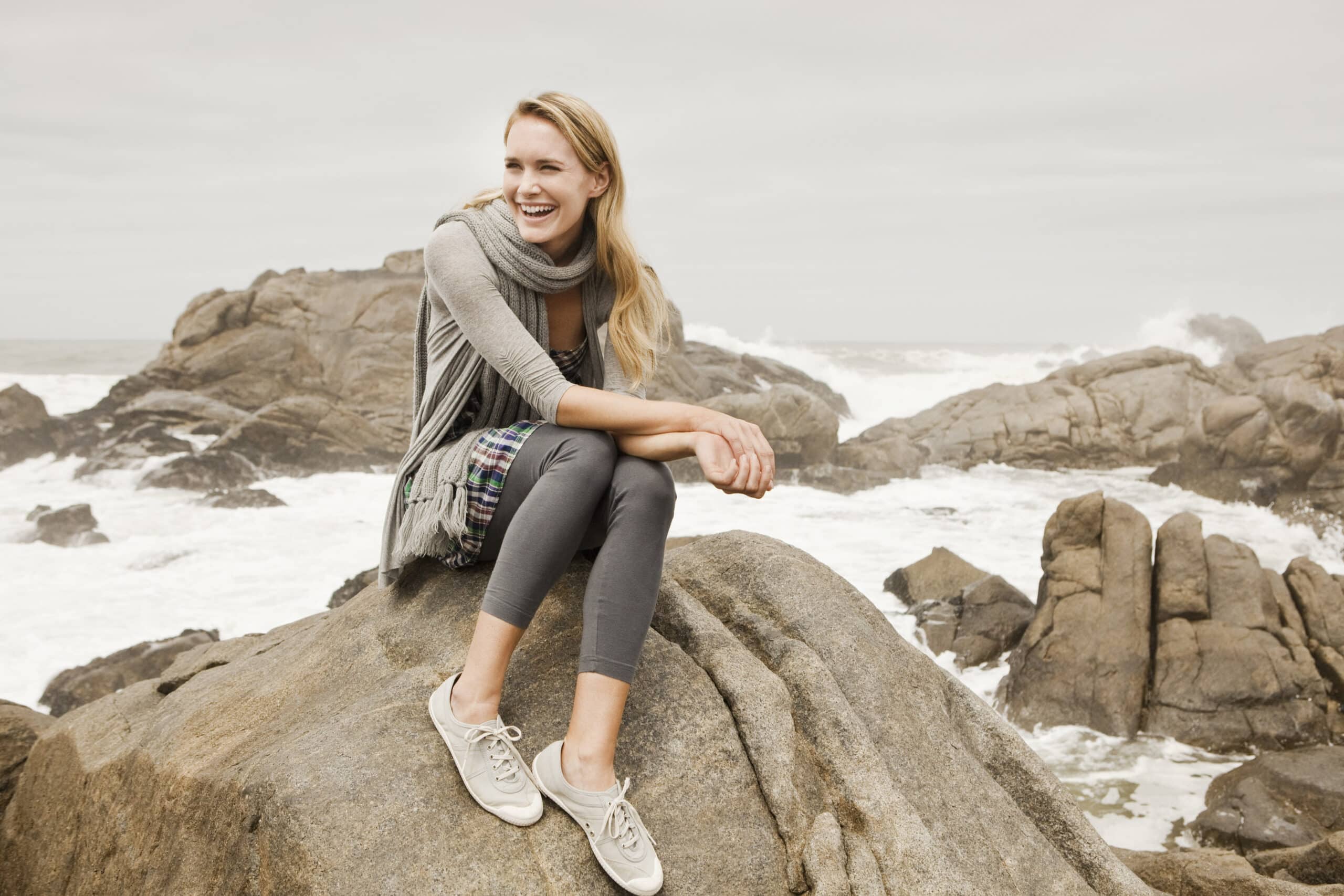 Eine fröhliche Frau sitzt lachend an einer felsigen Küste, im Hintergrund rauschen die Meereswellen. Sie trägt einen Schal, Leggings und Turnschuhe und hat eine Tasche über der Schulter. © Fotografie Tomas Rodriguez
