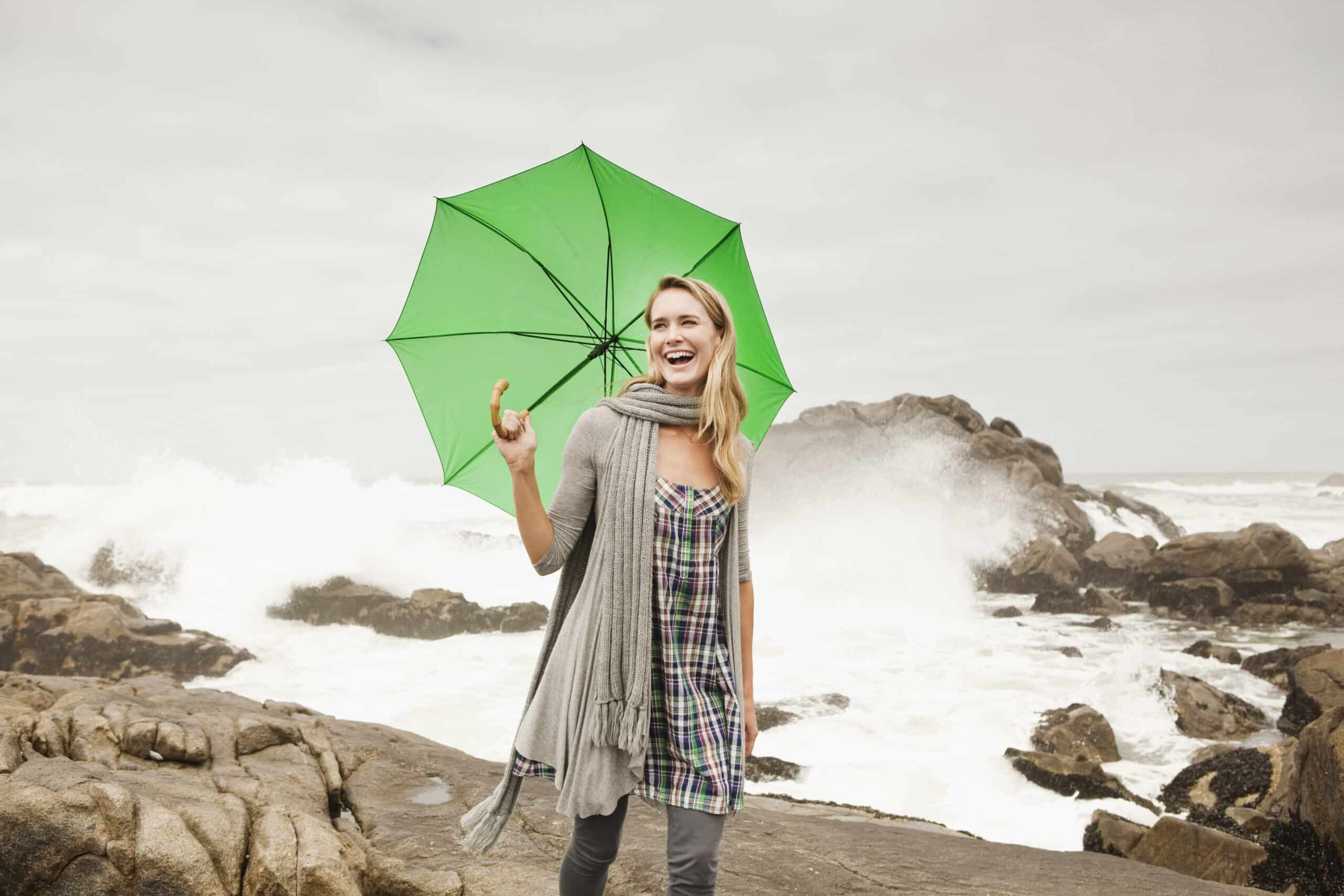 Eine fröhliche Frau mit einem grünen Regenschirm steht an einer felsigen Küste, hinter ihr brechen die Wellen. Sie trägt ein Kopftuch, lächelt breit und zeigt nach oben. © Fotografie Tomas Rodriguez