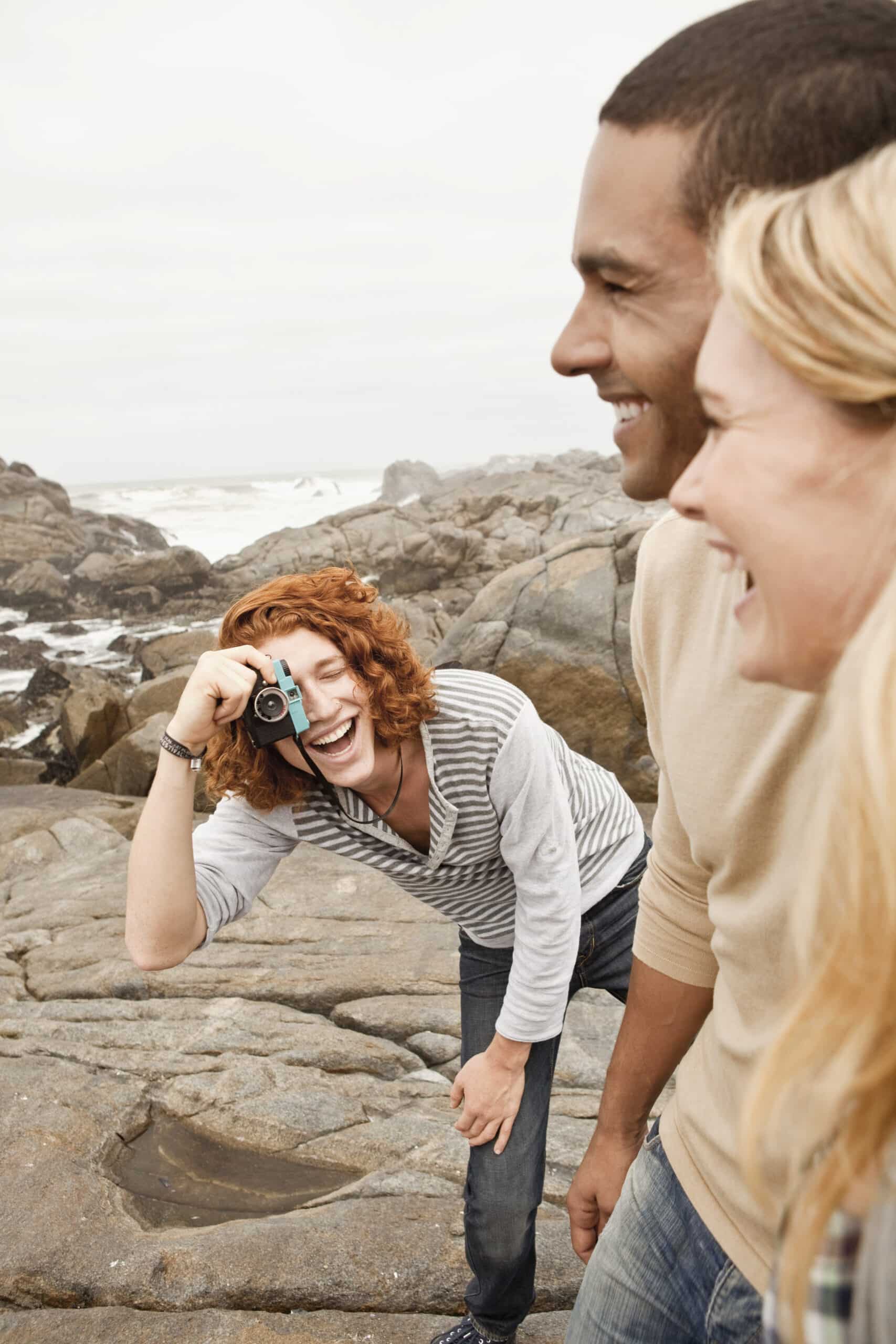 Eine fröhliche Frau mit lockigem rotem Haar macht mit einer Kamera ein Foto und lacht mit einem Mann und einer anderen Frau in einer felsigen Küstenlandschaft. © Fotografie Tomas Rodriguez