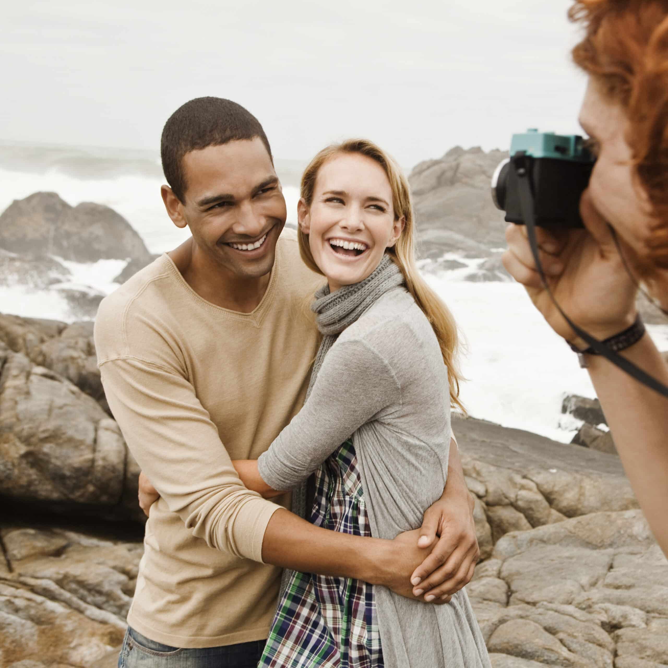 Ein fröhliches Paar umarmt sich an einer felsigen Küste, während eine Person ein Foto von ihnen macht. Der Mann, der ein hellbraunes Hemd trägt, lächelt die Frau an, die einen grauen Schal trägt, und beide strahlen Glück aus. © Fotografie Tomas Rodriguez