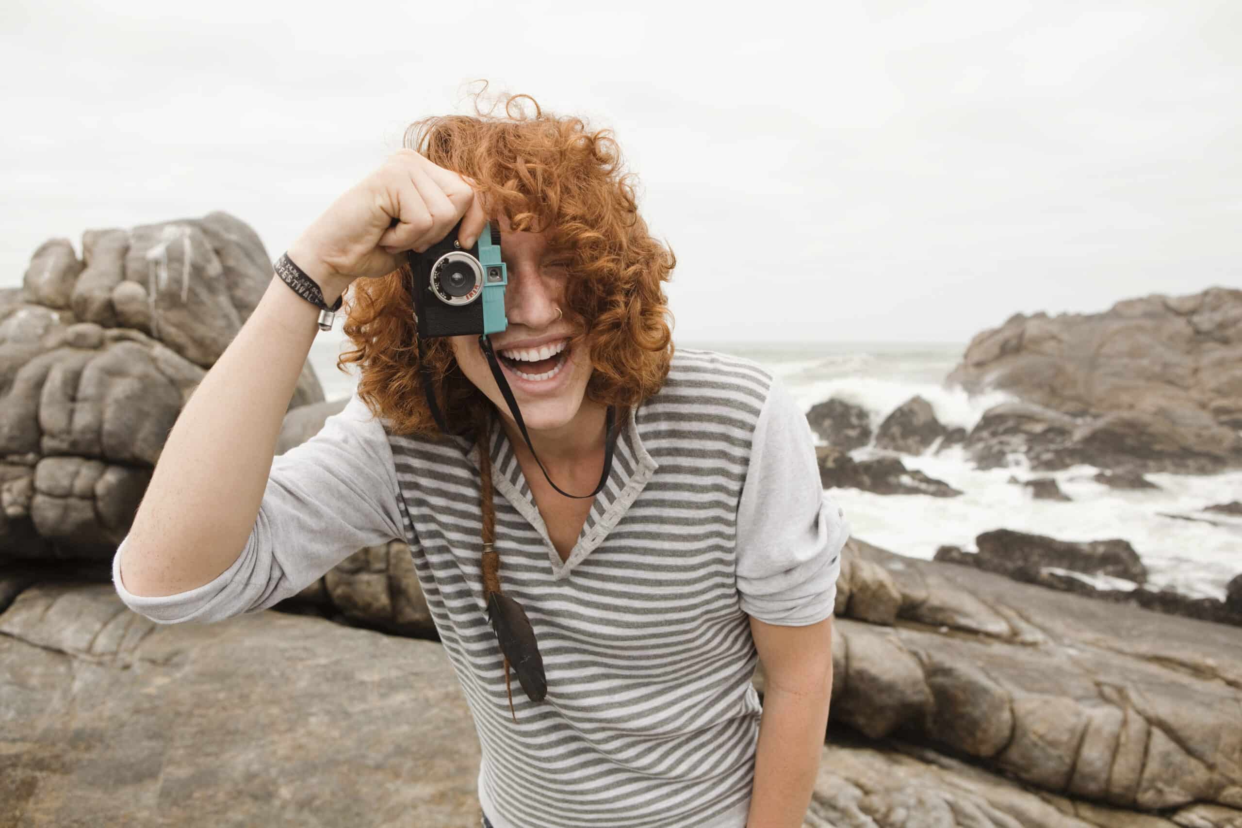 Eine fröhliche Frau mit lockigem rotem Haar macht mit einer Vintage-Kamera ein Foto und steht auf felsigem Küstengelände vor einem bewölkten Himmel. © Fotografie Tomas Rodriguez