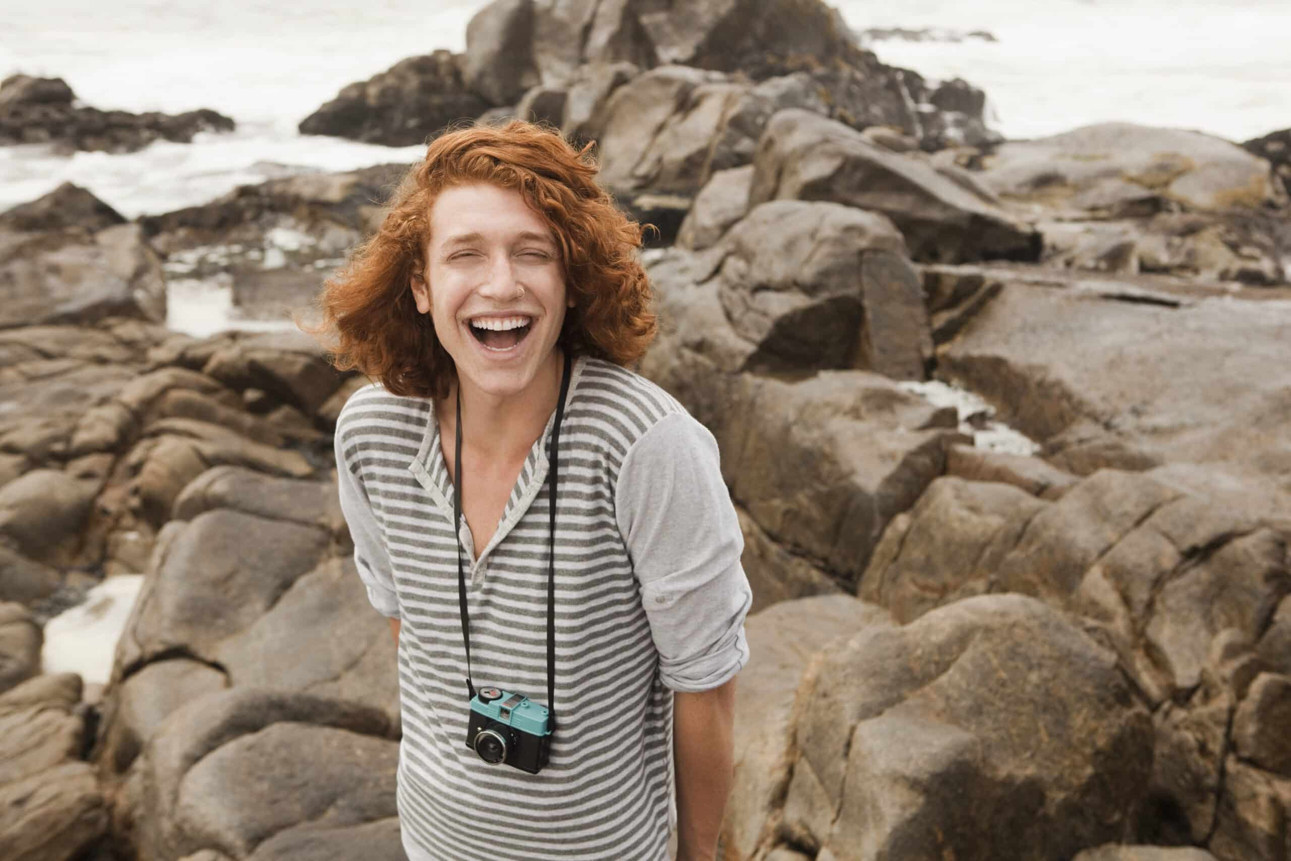 Ein fröhlicher junger Mensch mit lockigem roten Haar und einer Kamera um den Hals lacht herzlich an einem steinigen Strand mit Meereswellen im Hintergrund. © Fotografie Tomas Rodriguez