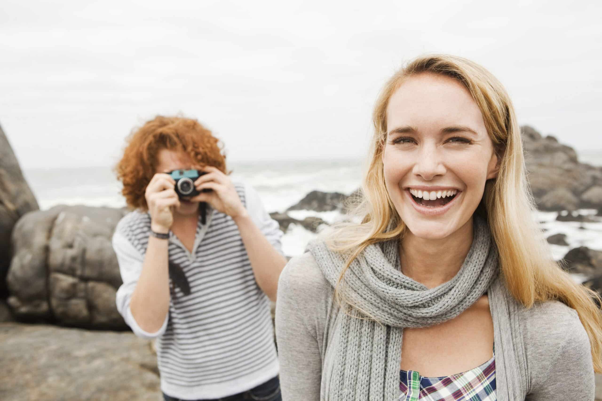 Eine fröhliche Frau mit blondem Haar lächelt in die Kamera und trägt ein Kopftuch. Im Hintergrund macht eine Person mit lockigem Haar ein Foto von ihr an der felsigen Küste. © Fotografie Tomas Rodriguez