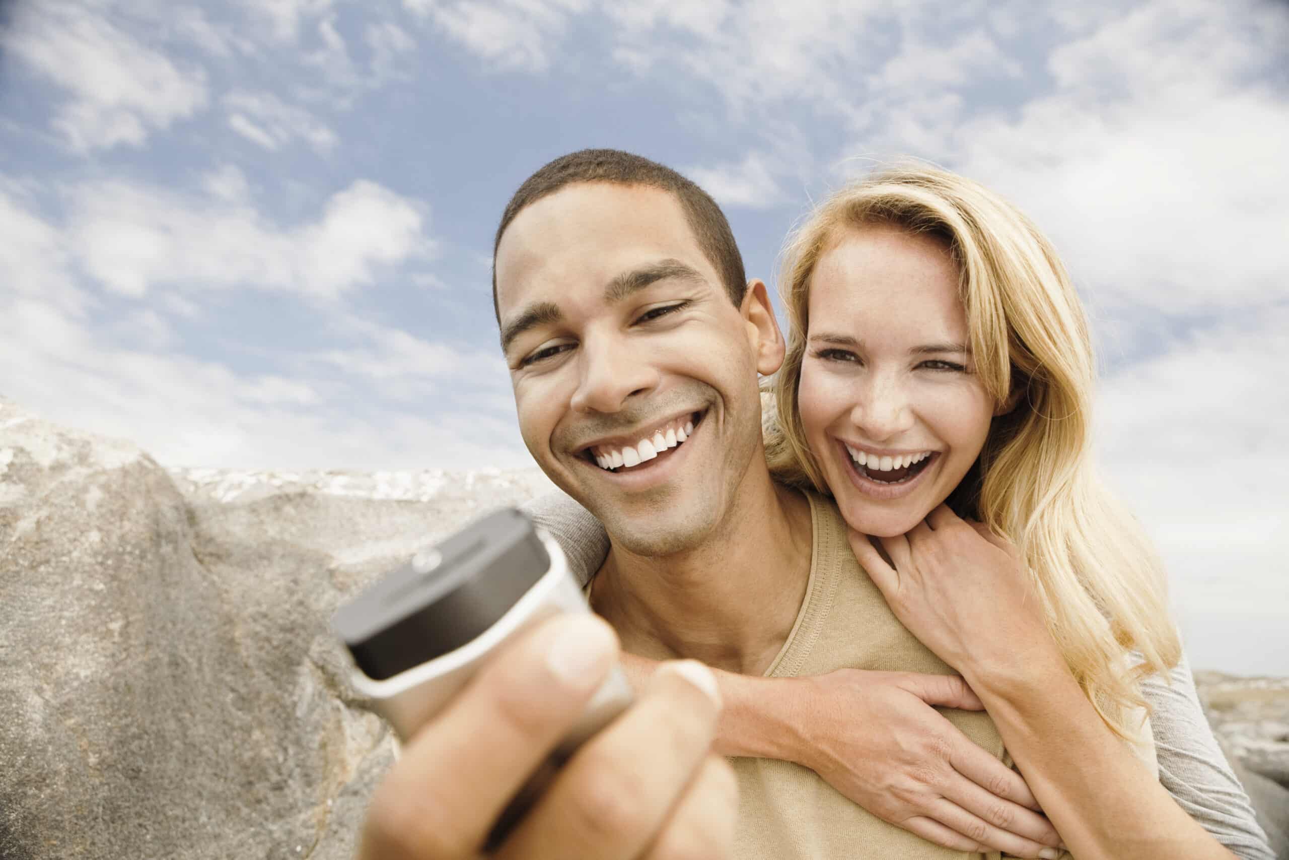 Ein fröhliches Paar macht im Freien ein Selfie. Der Mann hält die Kamera und beide lächeln breit vor dem Hintergrund eines bewölkten Himmels und eines felsigen Geländes. © Fotografie Tomas Rodriguez