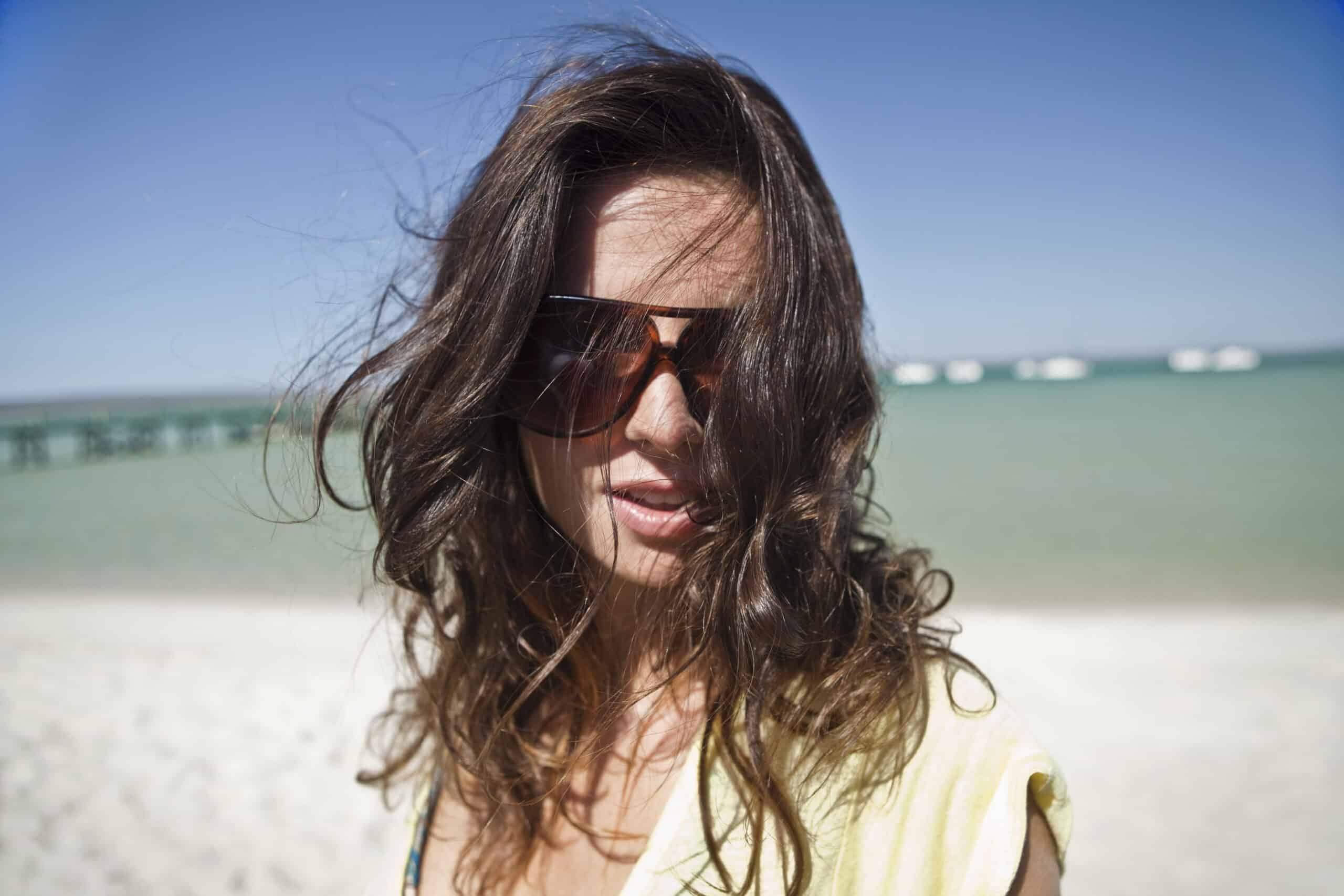 Eine Frau mit zerzaustem Haar und Sonnenbrille steht an einem sonnigen Strand. Im Hintergrund ist das Meer zu sehen. Ihr Gesichtsausdruck ist verschwommen, wirkt aber fröhlich. © Fotografie Tomas Rodriguez
