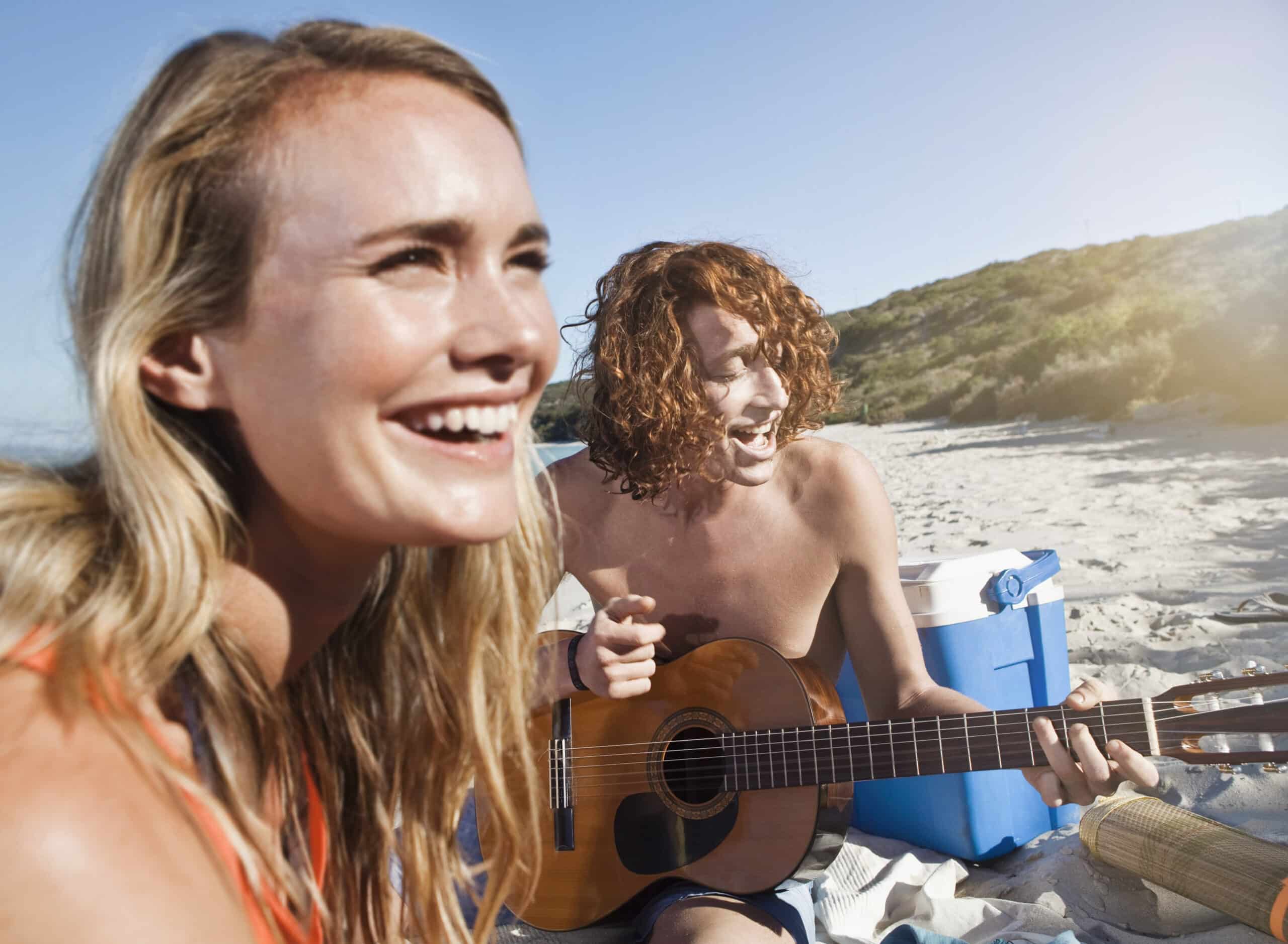 Zwei fröhliche Menschen an einem sonnigen Strand, einer spielt Gitarre, der andere lacht, mit einer Kühlbox in der Nähe, und genießen einen verspielten, musikalischen Strandtag. © Fotografie Tomas Rodriguez